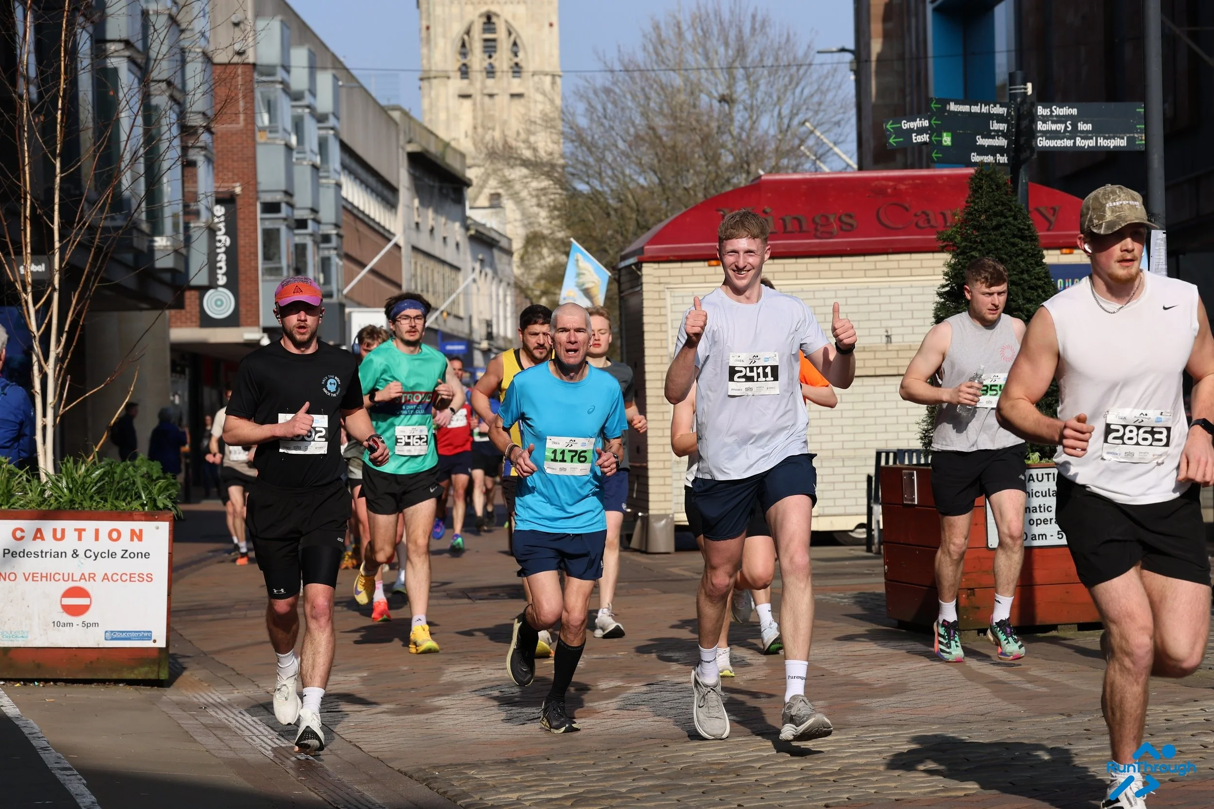 A large group of runners, headed up by around 10 people, including a man running with his thumbs up. Gloucester Cathedral is in the background and the runners are on a pedestrianised section of the course.