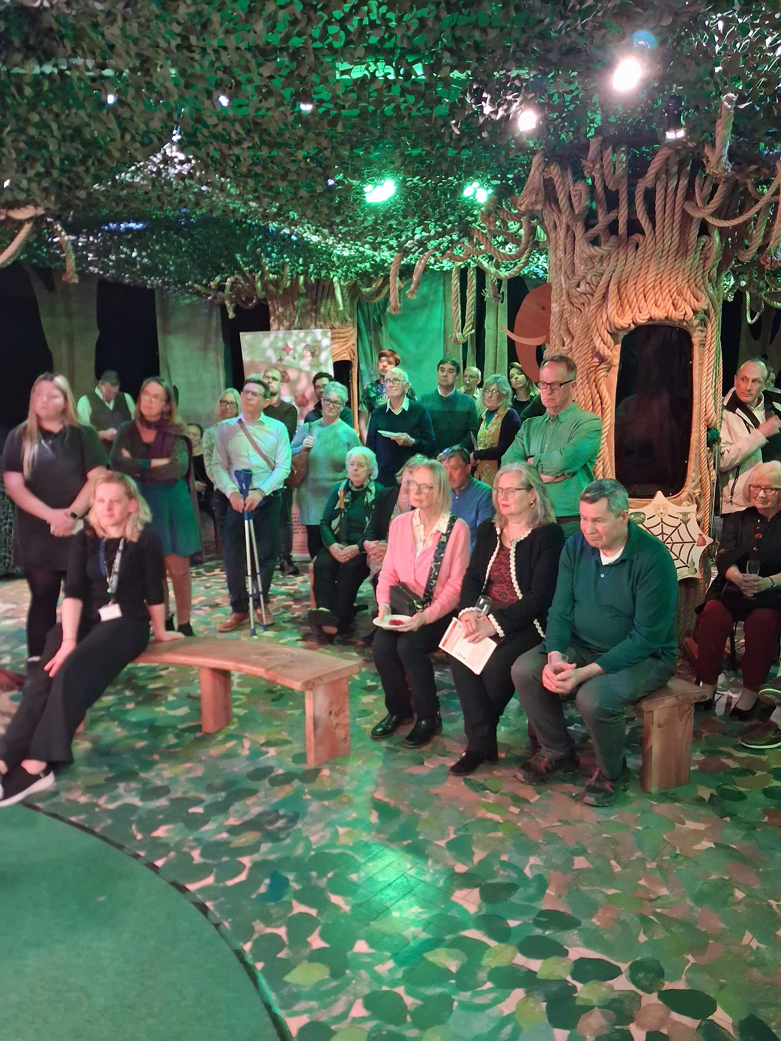 A large group of around 25 people, sitting and standing beneath a leafy canopy and trees made out of rope.