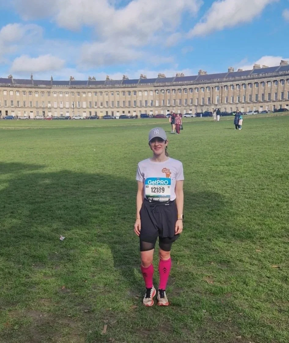 An African Children's Fund fundraiser, wearing a charity t-shirt with the logo on the front and with a race number pinned on. She's standing in front of Bath's famous Royal Crescent.