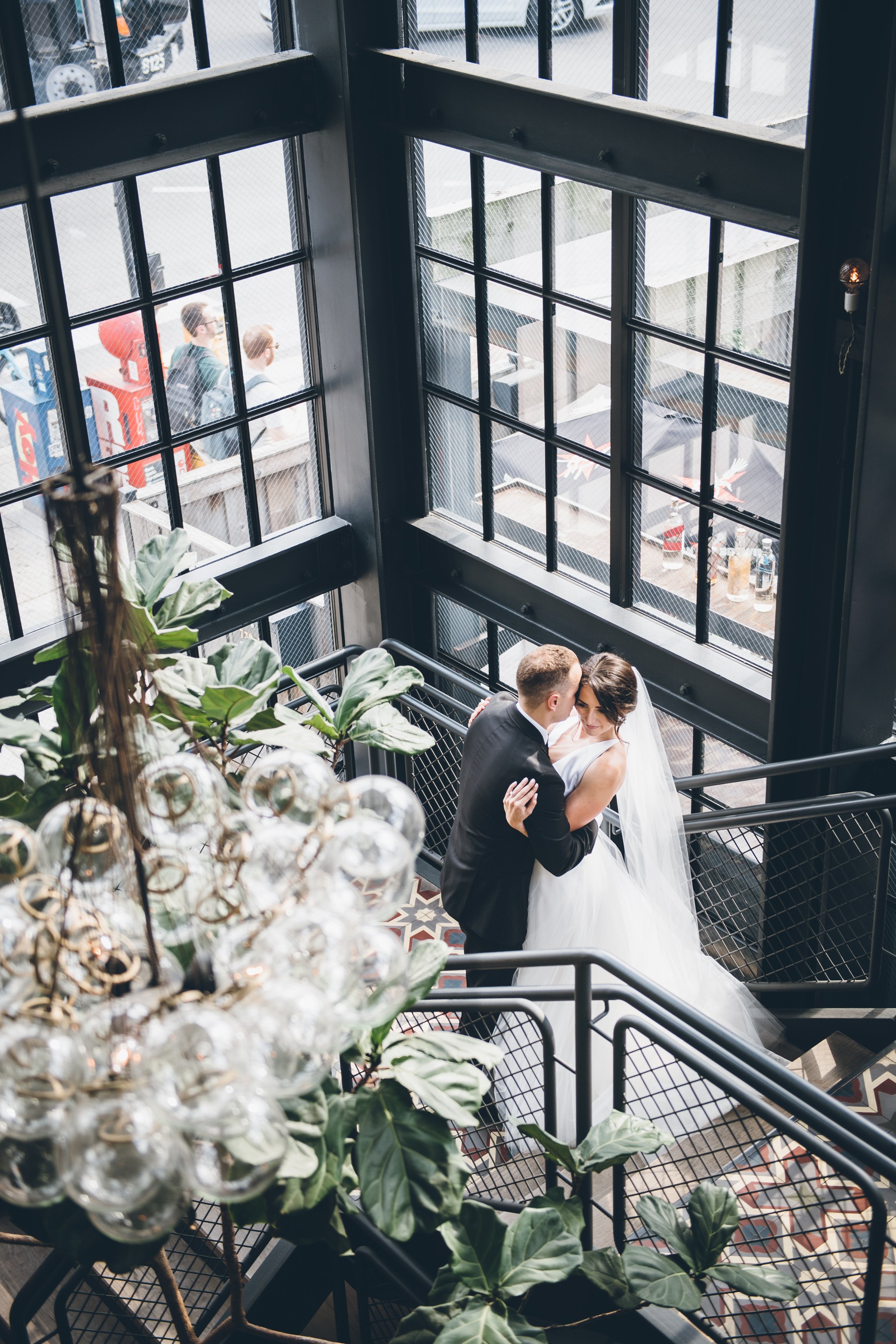 A bride and groom dancing on a staircase inside a modern building with large grid windows, plants, and a chandelier in the foreground, with people visible outside the window.