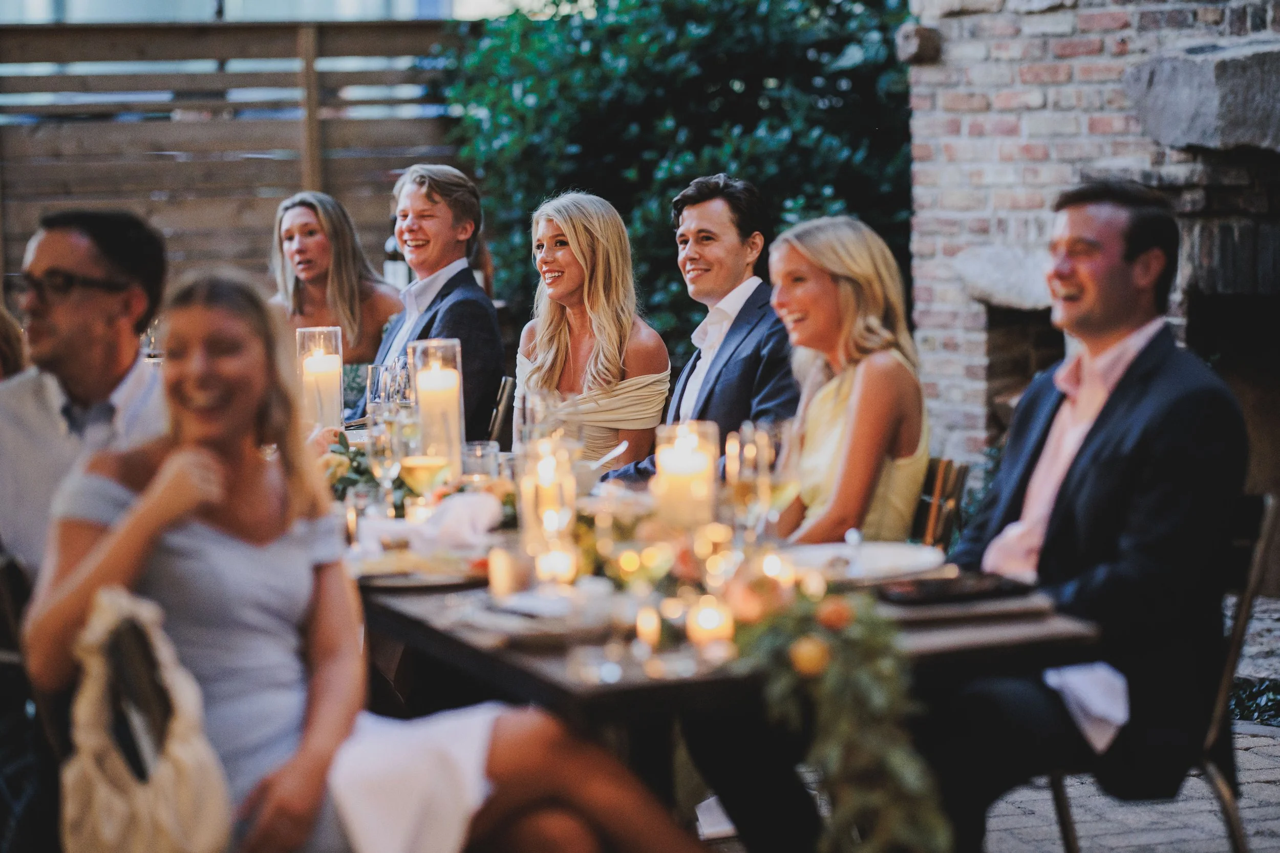 A group of friends or family sitting at a dinner table during an outdoor evening party, with candles and floral decorations on the table, surrounded by a brick wall and wooden fencing.