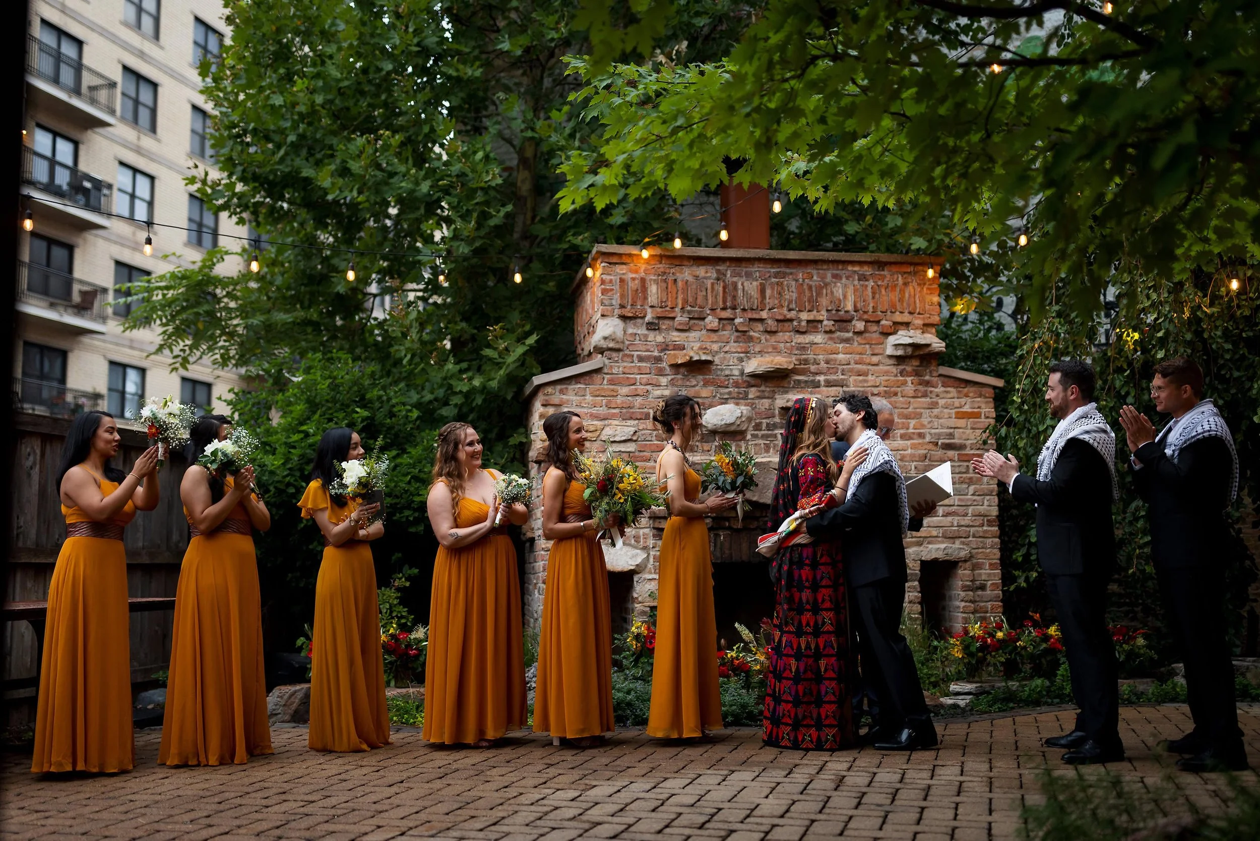 A same-sex couple is sharing a kiss during a wedding ceremony, surrounded by bridesmaids and groomsmen, outdoors under string lights, with a brick fireplace and greenery in the background.