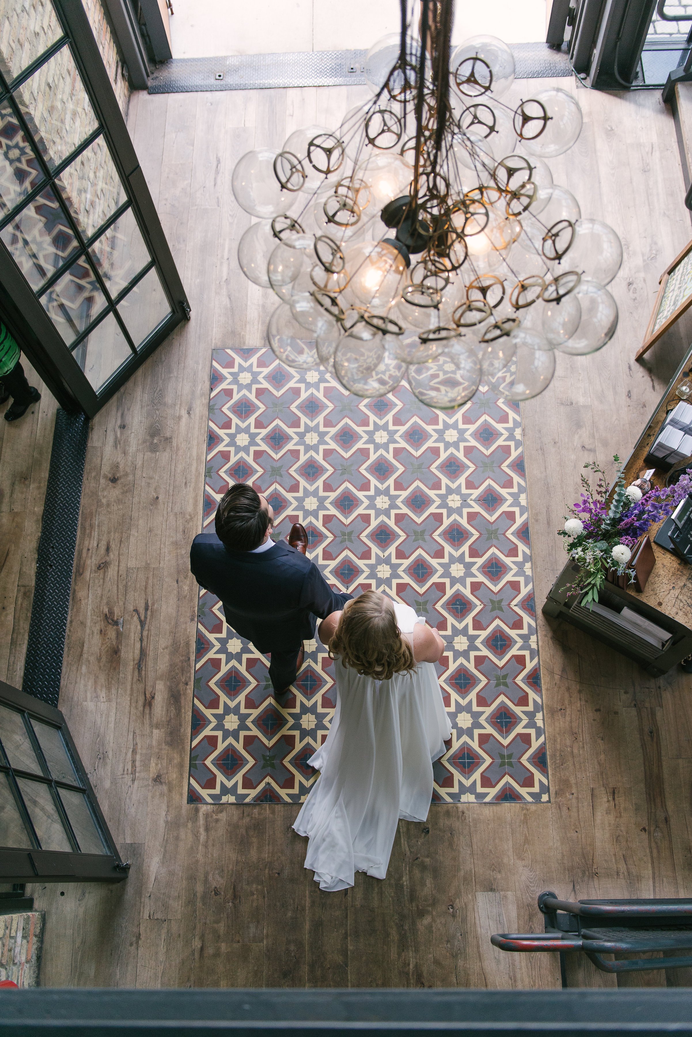 A bride and groom walking together on a patterned rug inside a building, viewed from above. A chandelier with exposed bulbs and decorative loops hangs overhead. The entrance features glass doors, and there are flowers on a counter to the right.