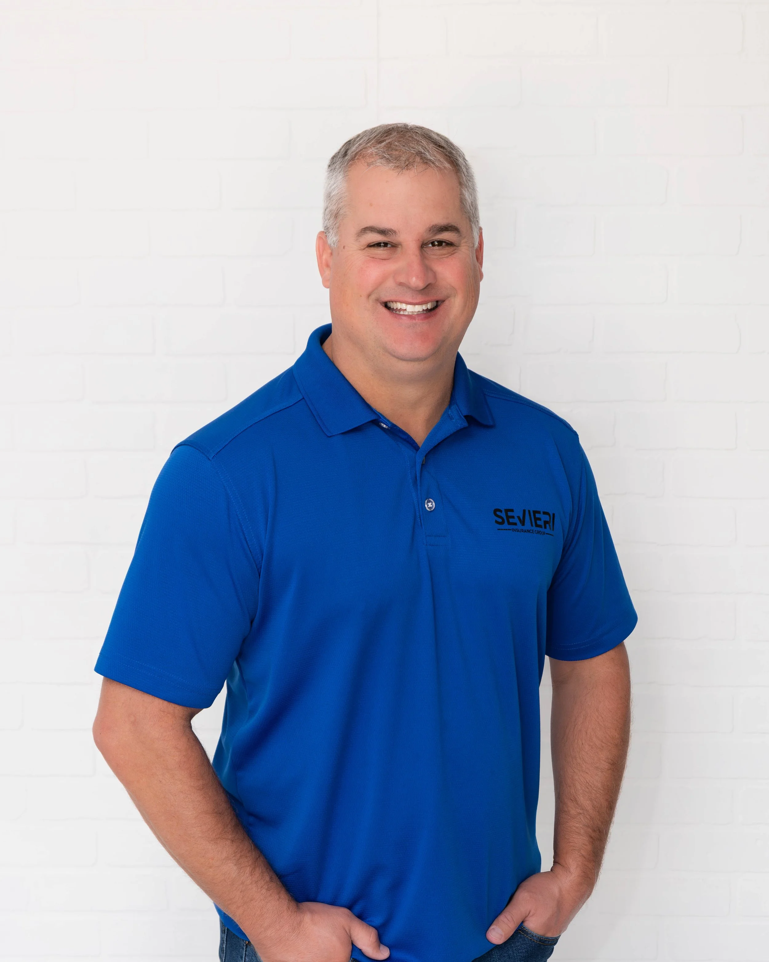 A smiling man wearing a blue polo shirt with the logo of an insurance company, standing in front of a white brick wall.