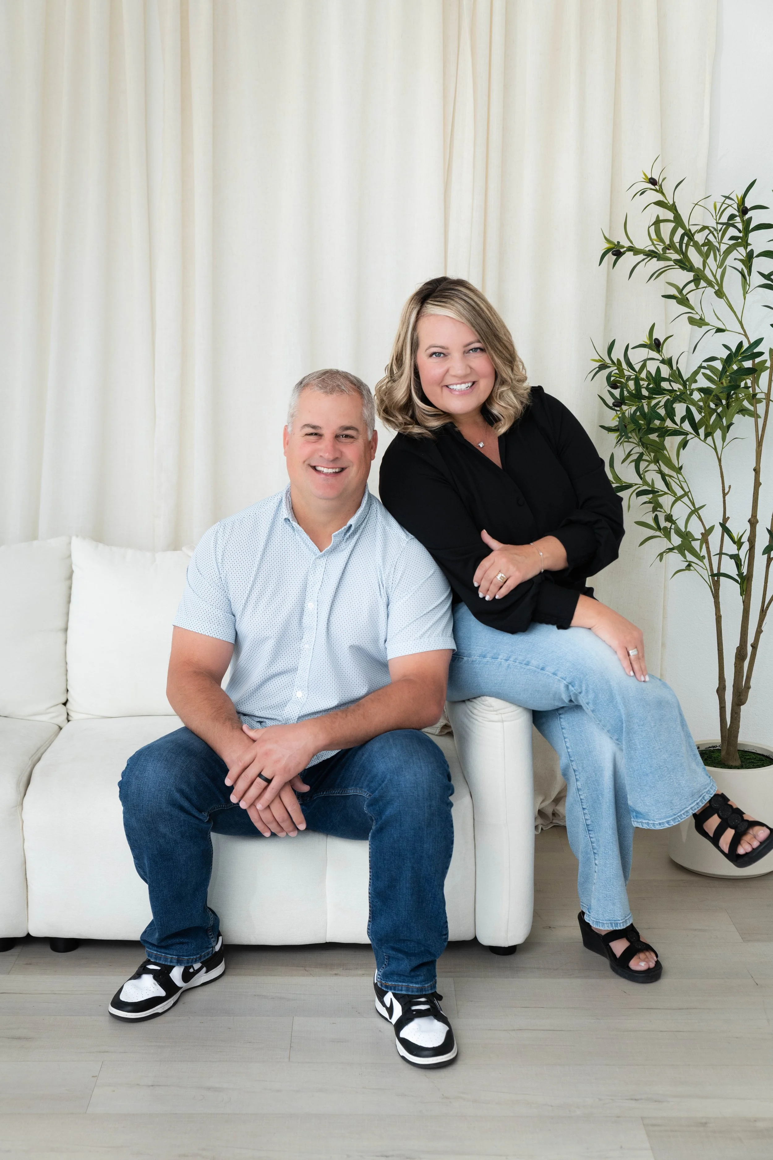 Two smiling adults, a man and a woman, sitting on a white couch in a room with a white curtain backdrop and a potted plant on the right.