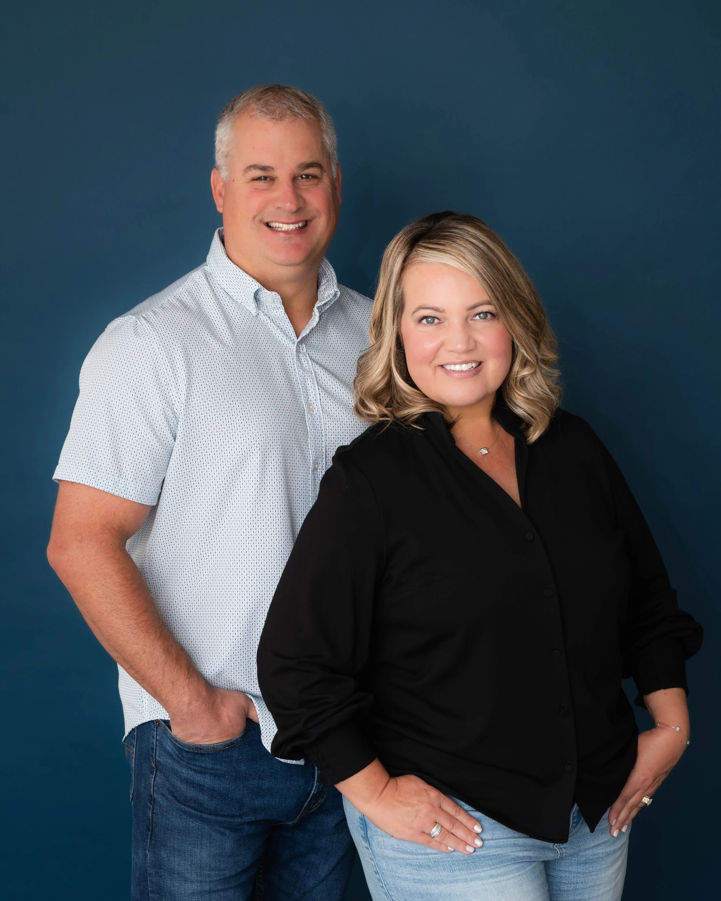 A smiling man in a white patterned shirt and blue jeans standing next to a woman in a black shirt and light blue jeans against a dark blue background.