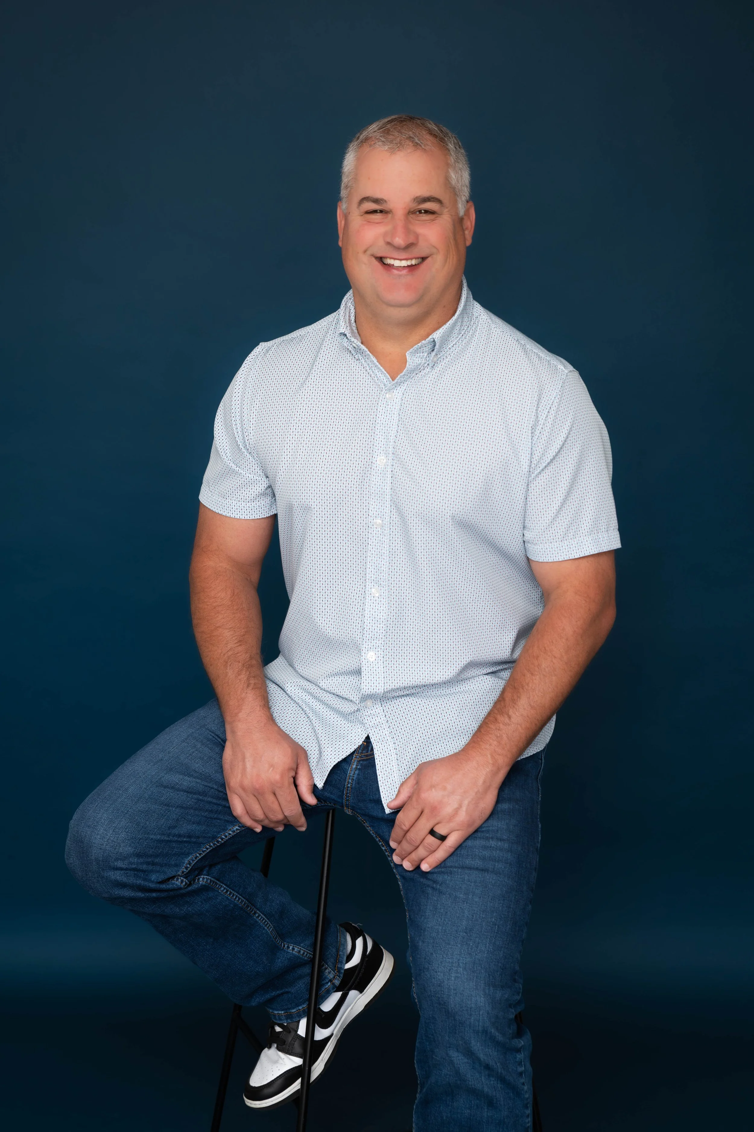A man with short gray hair and a wide smile sitting on a stool against a dark blue background. He is wearing a light-colored, short-sleeved button-up shirt with small patterns, blue jeans, and black and white sneakers.