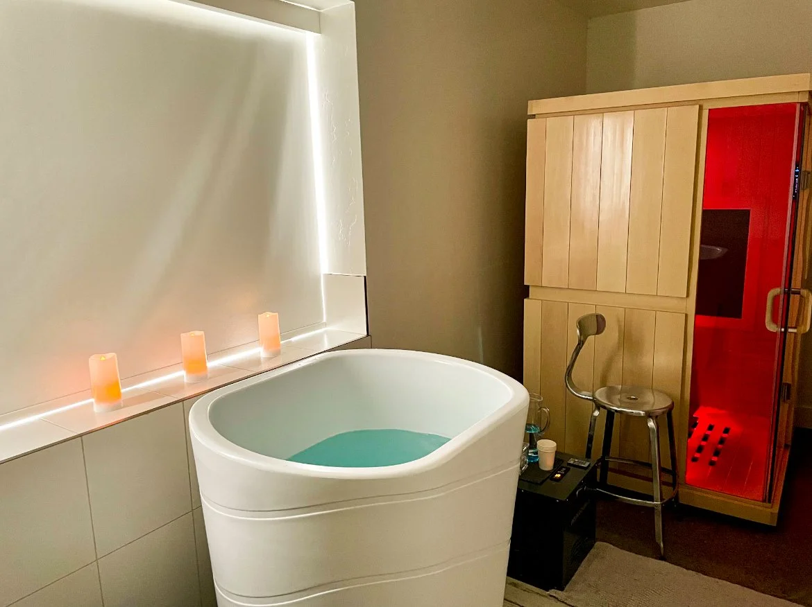 A modern sauna room with a white cold plunge filled with water, three candles on the ledge, a wooden sauna, a metal stool, and a red light in the sauna.