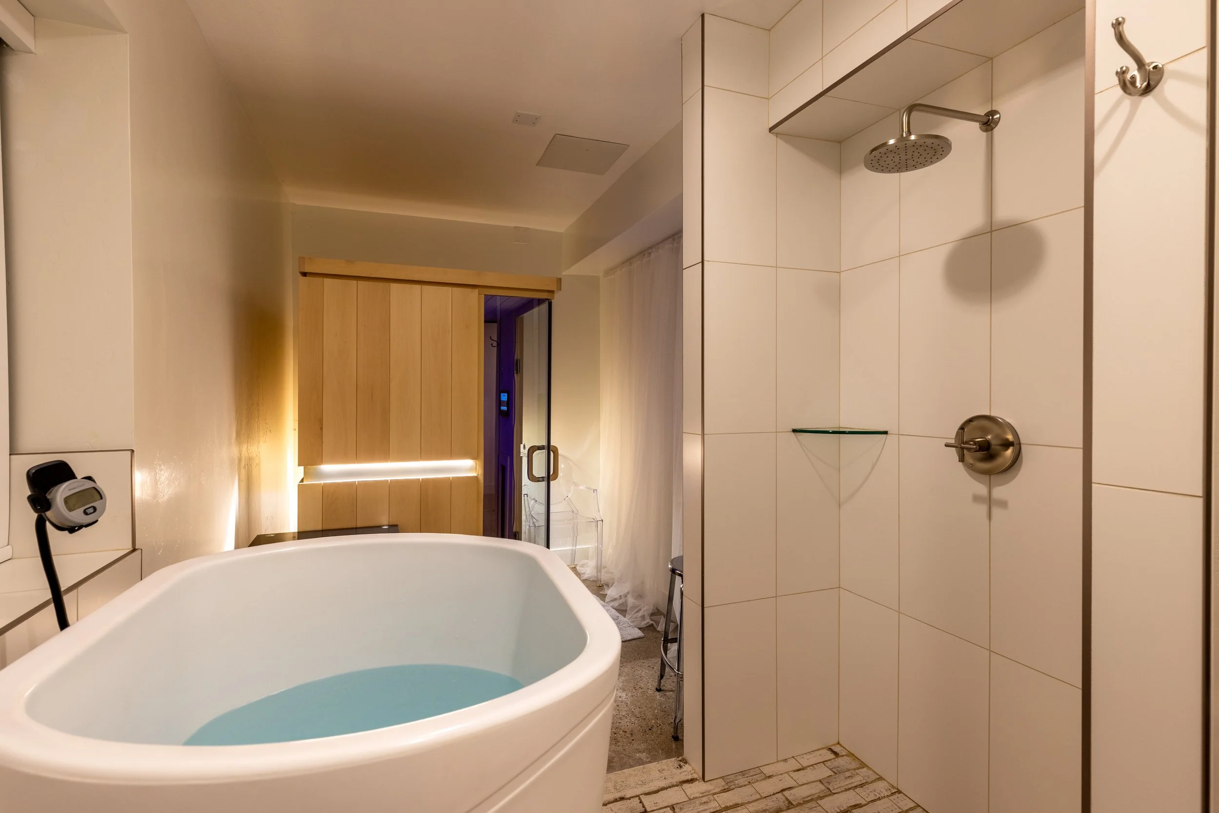 Sauna room with a white cold plunge filled with water, presenting a shower area with white tiled walls, a ceiling showerhead, and a warm wooden sauna at the back.
