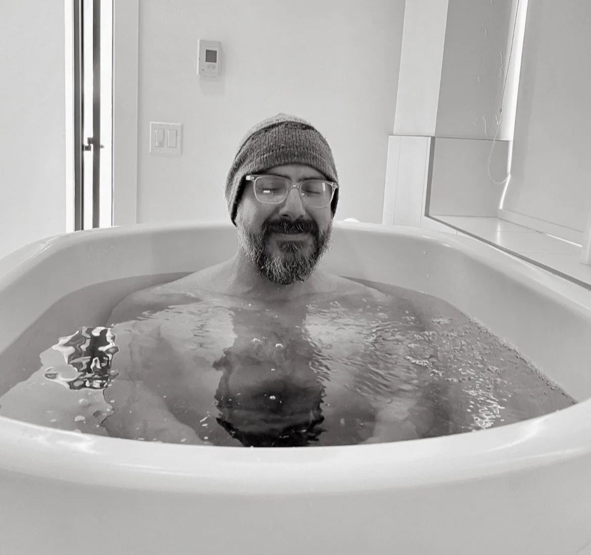 A man with glasses and a beard relaxing in a cold plunge, wearing a beanie.