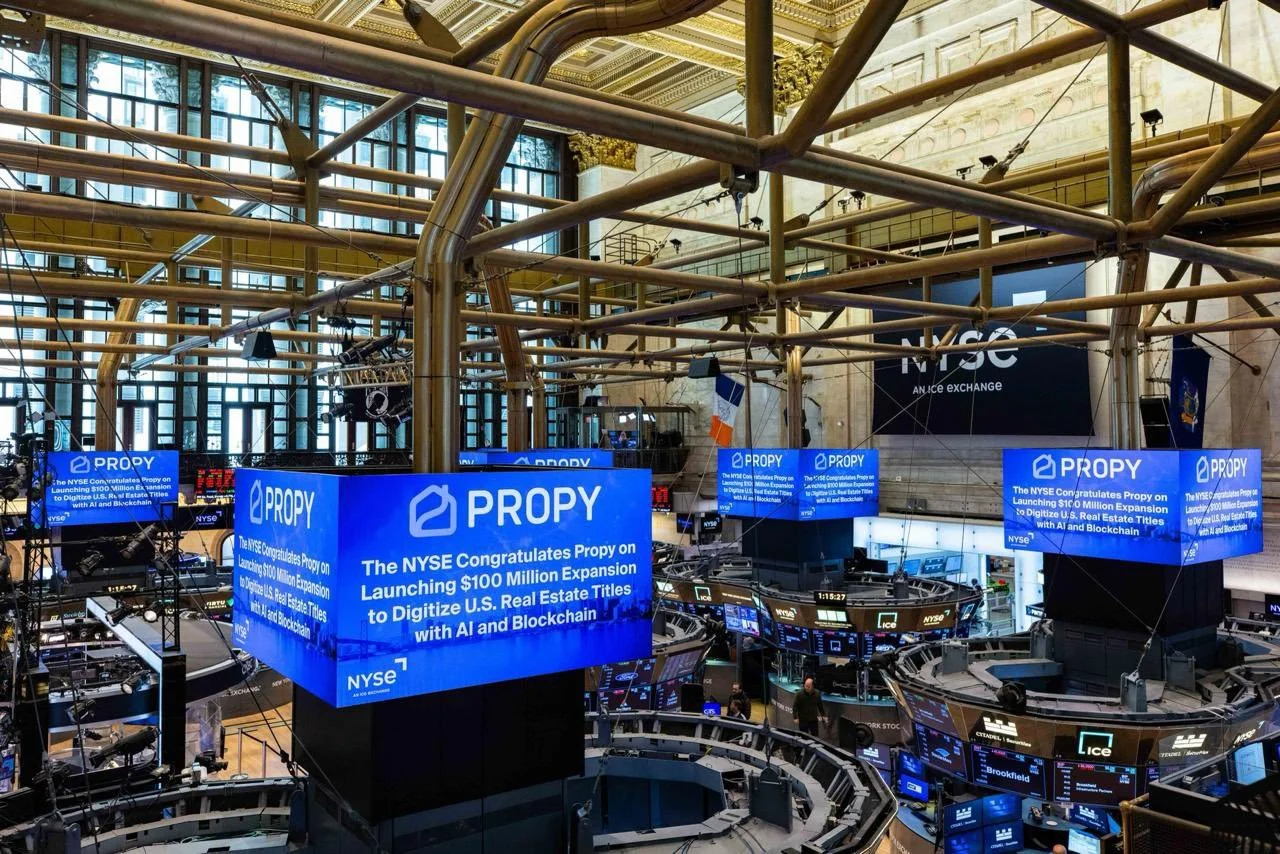 The New York Stock Exchange trading floor with multiple large blue digital screens displaying a message about Propy's $100 million expansion launch for digitizing U.S. real estate titles using AI and blockchain, with traders and monitors in the background.