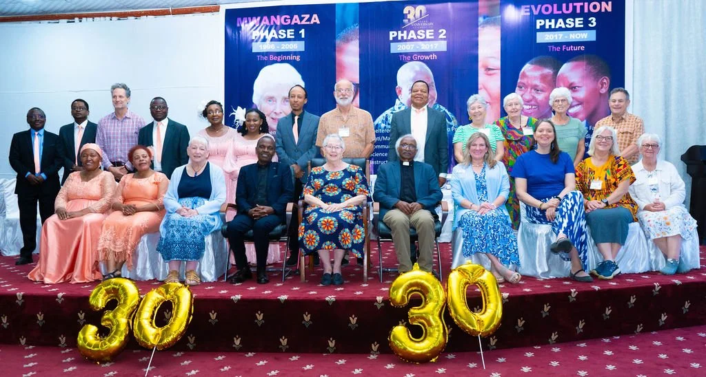 Tanzanians and Americans sit on a stage together, smiling for a group photo. The background includes a photo of Mama Shoonie Hartwig. Gold balloons in the shape of the number 30 are in the foreground.