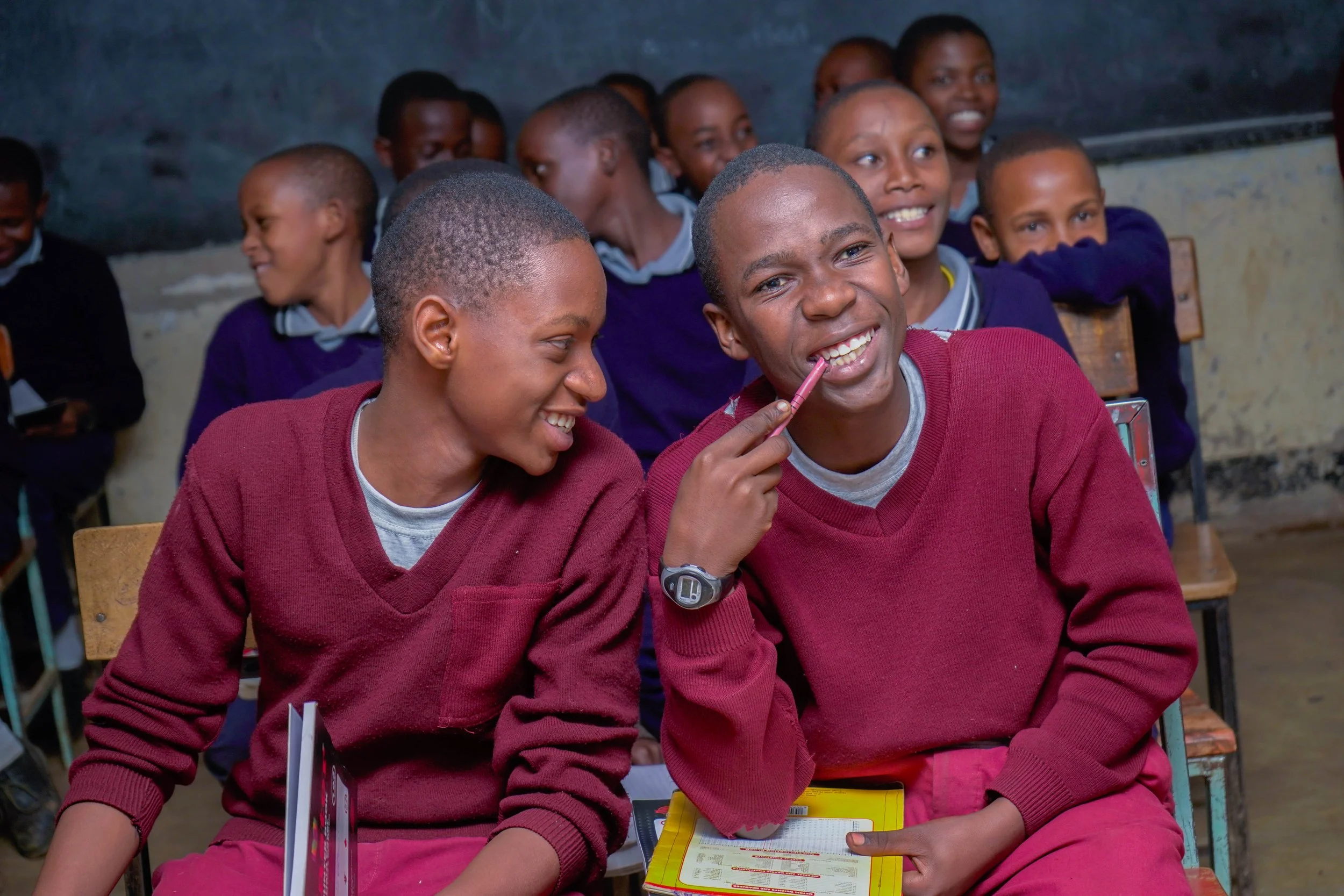 Two Tanzanian school boys sit together at a desk laughing.