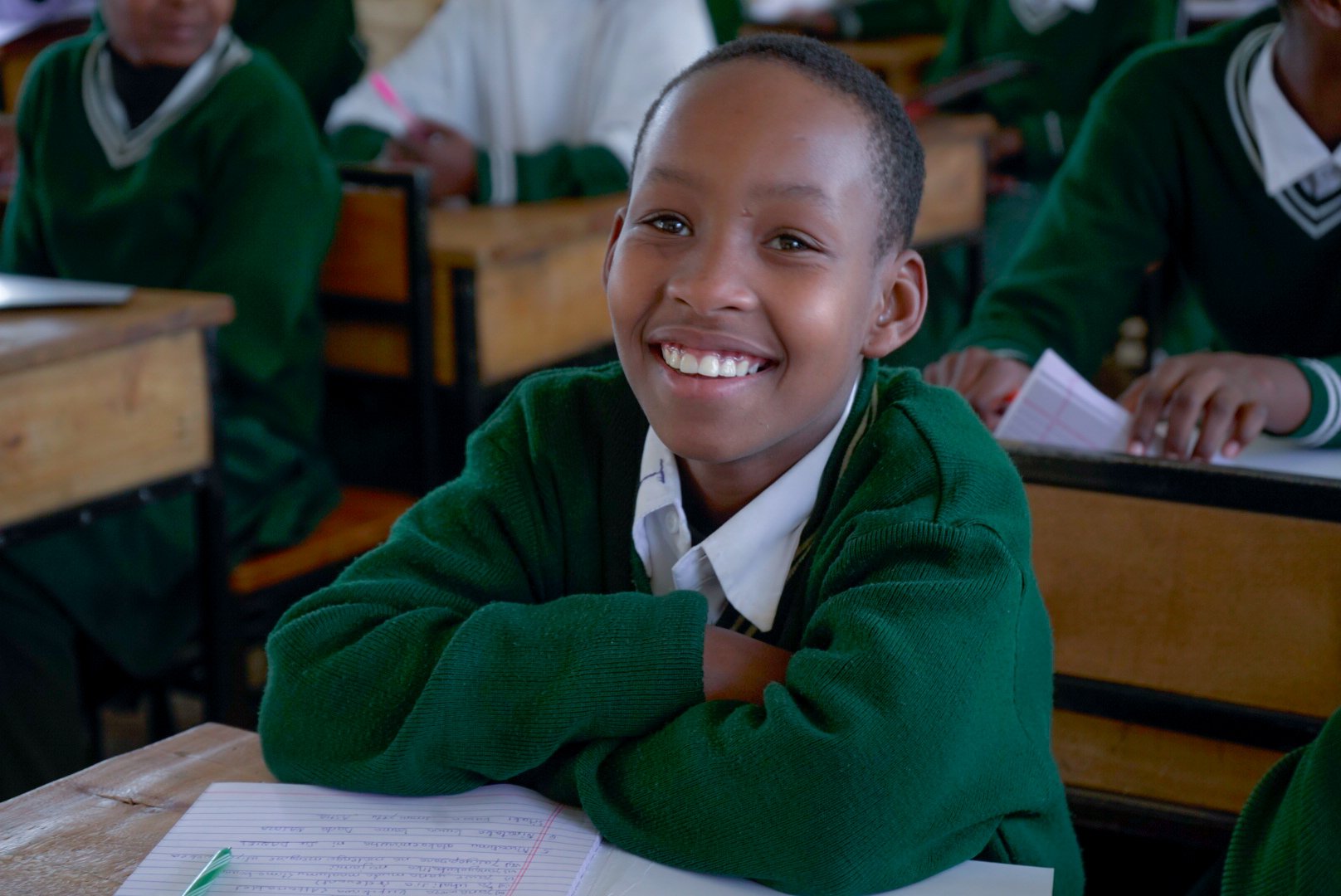 A Tanzanian secondary student smiles broadly for the camera. She has short black hair, brown eyes, and a green sweater.