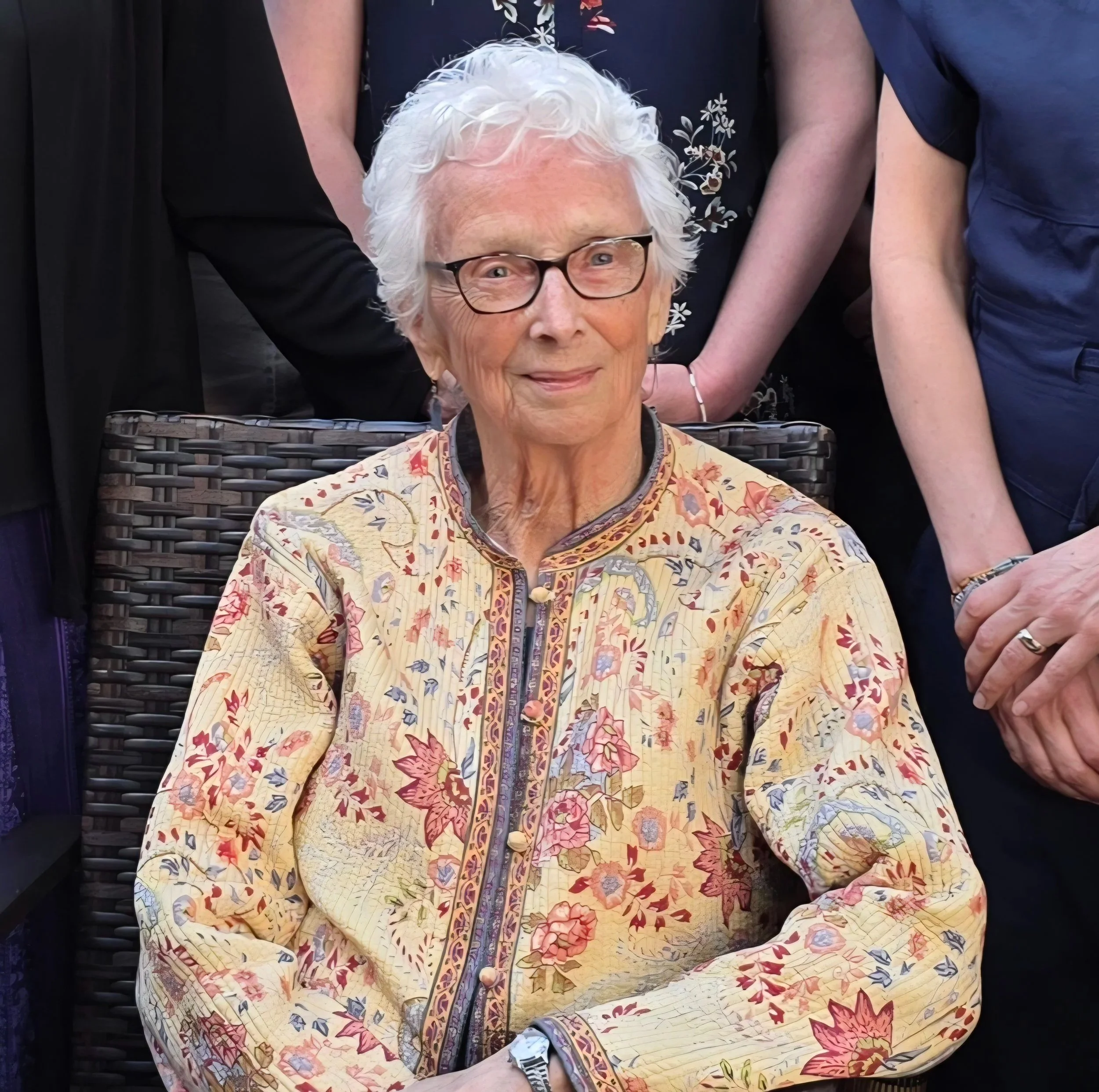 Shoonie is an older white woman with short, wavy, white hair. She is wearing glasses and a yellow quilted jacket.