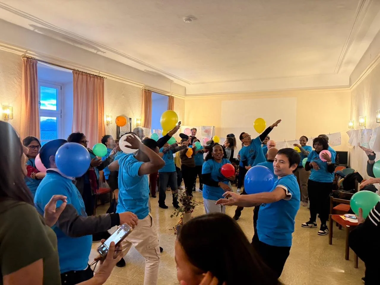 Participants in blue shirts play an NVC game with balloons. They are inside a large room in the nunnery.