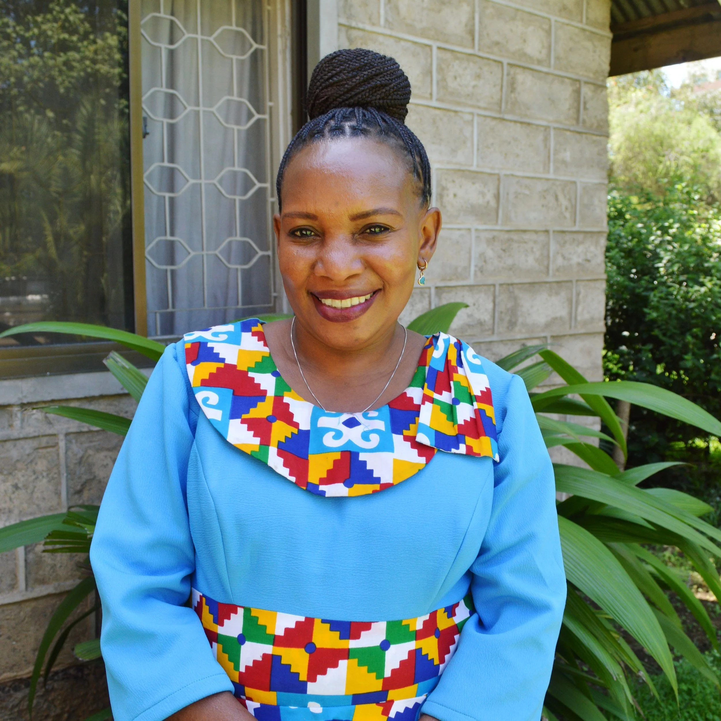 Salome is a Tanzanian woman. Her hair is in small braids and pulled up into a bun. She is wearing a bright blue dress with a multicolored collar.