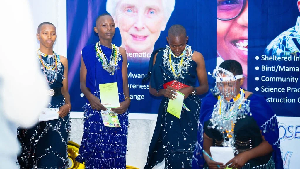 Female students perform a skit at the celebration. They wear blue robes with heavily beaded necklaces. One student wears a beaded headdress.
