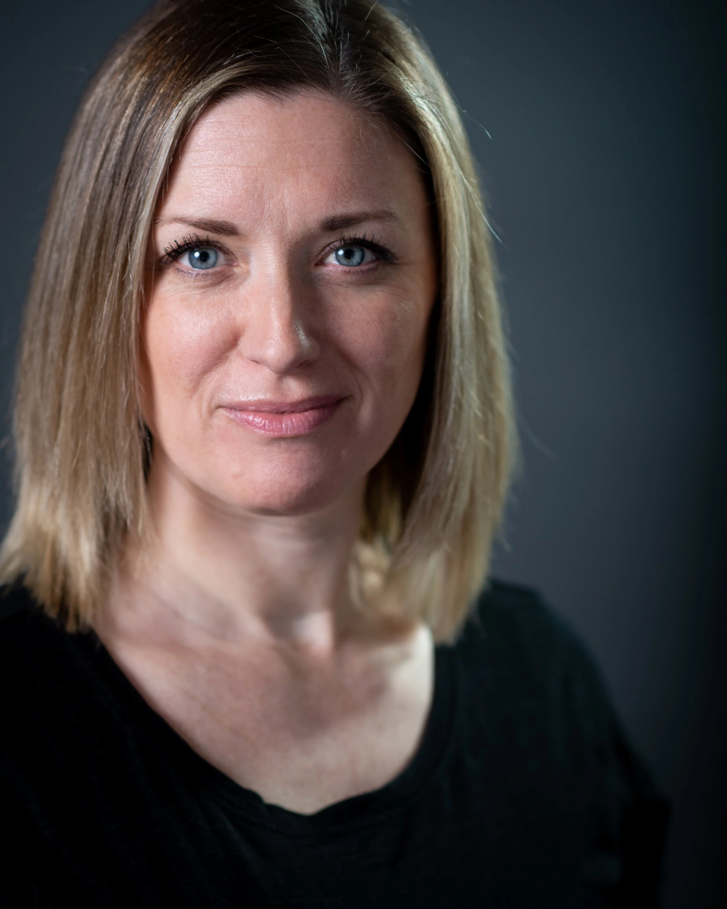 Gemma Boaden, headshot photo, with blonde hair, blue eyes, and fair skin, wearing a black top, against a dark background.