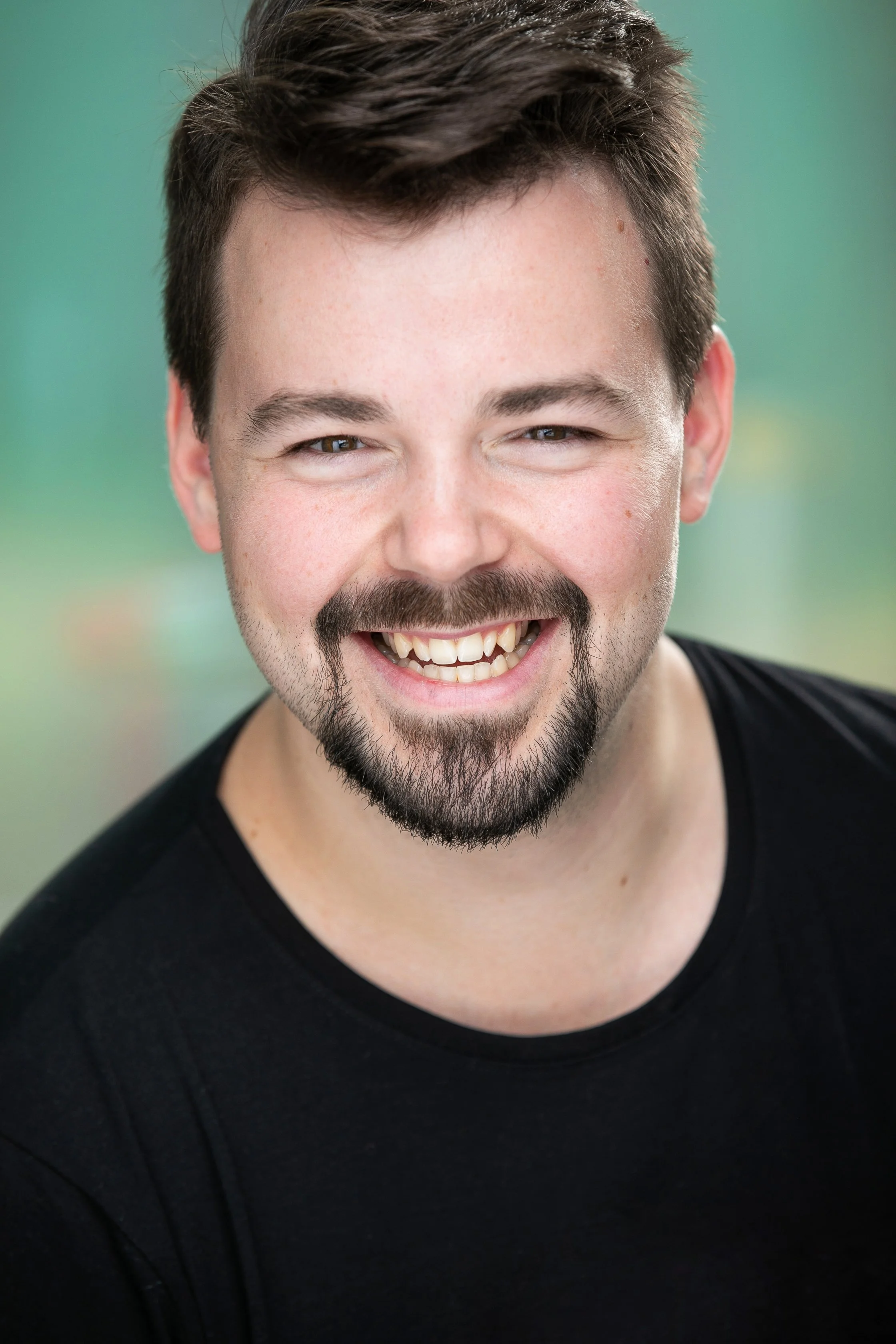 Wictor Koch, headshot photo, smiling with brown hair, a beard and mustache, wearing a black shirt, against a green blurred background.