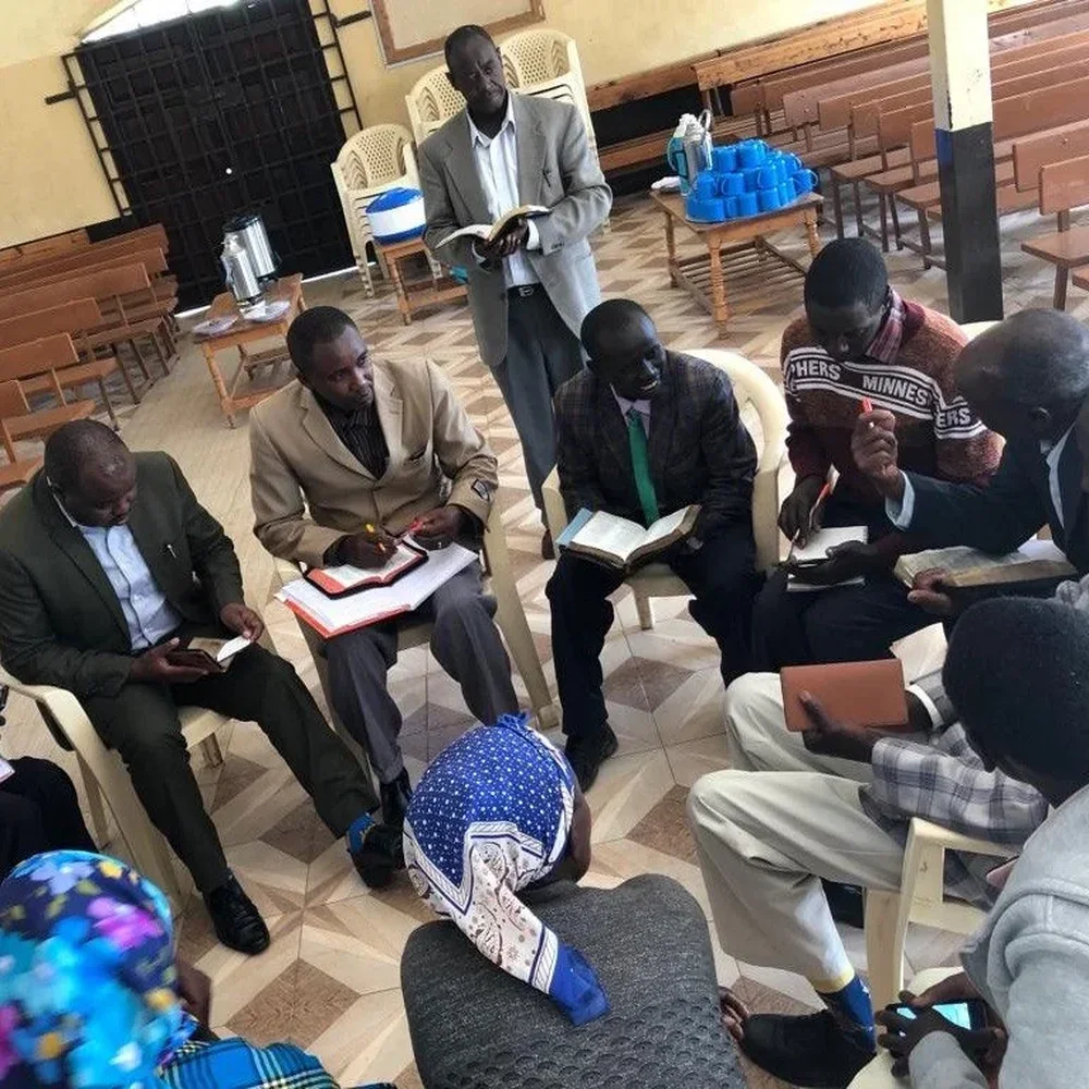 A group of pastors who are part of the pastors team from Kenya sitting in a circle, some holding bibles or phones, in a room with wooden flooring and chairs. Two men are standing, with one reading the bible aloud.