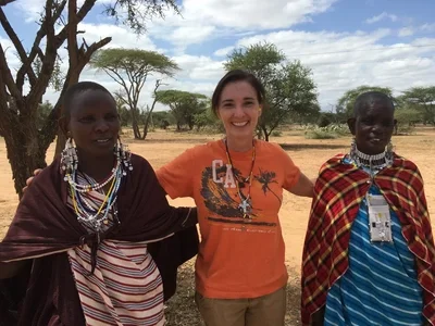 Tamara on her trip to Tanzania met two women standing outdoors under trees in a dry landscape, smiling, with blue sky and clouds above.