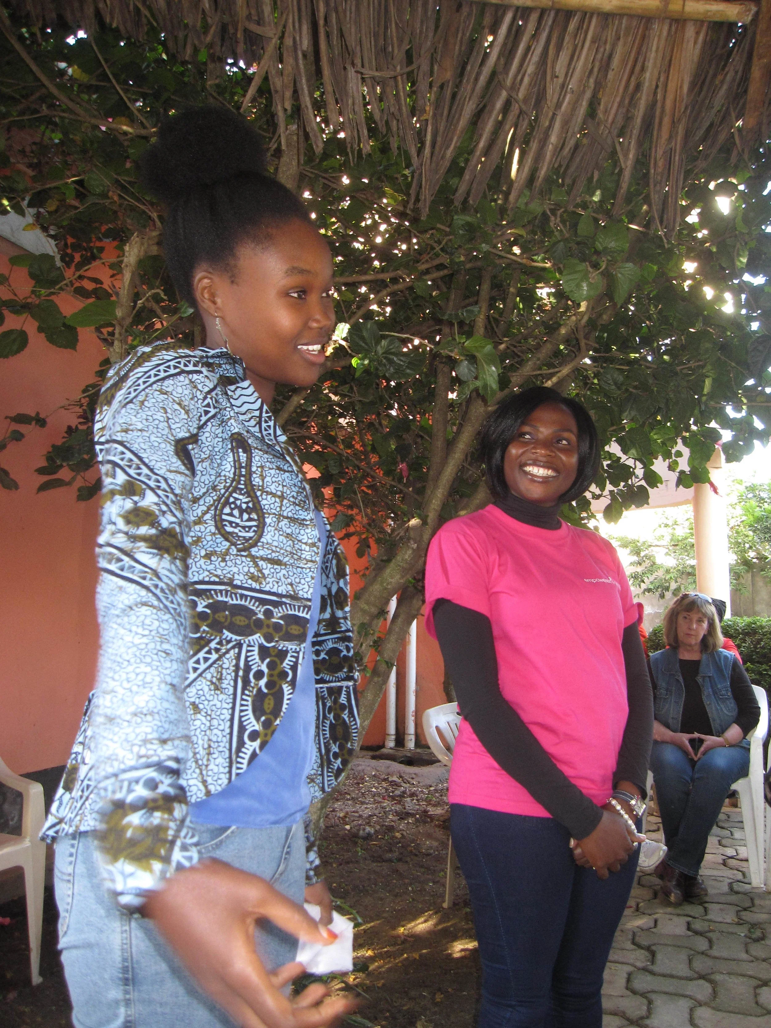 Two women standing near a tree, smiling and talking, with a woman seated in the background.