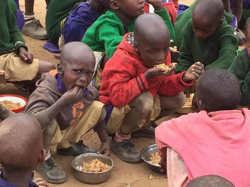 Lesing'ta Primary School children sitting on the ground, eating food from bowls.