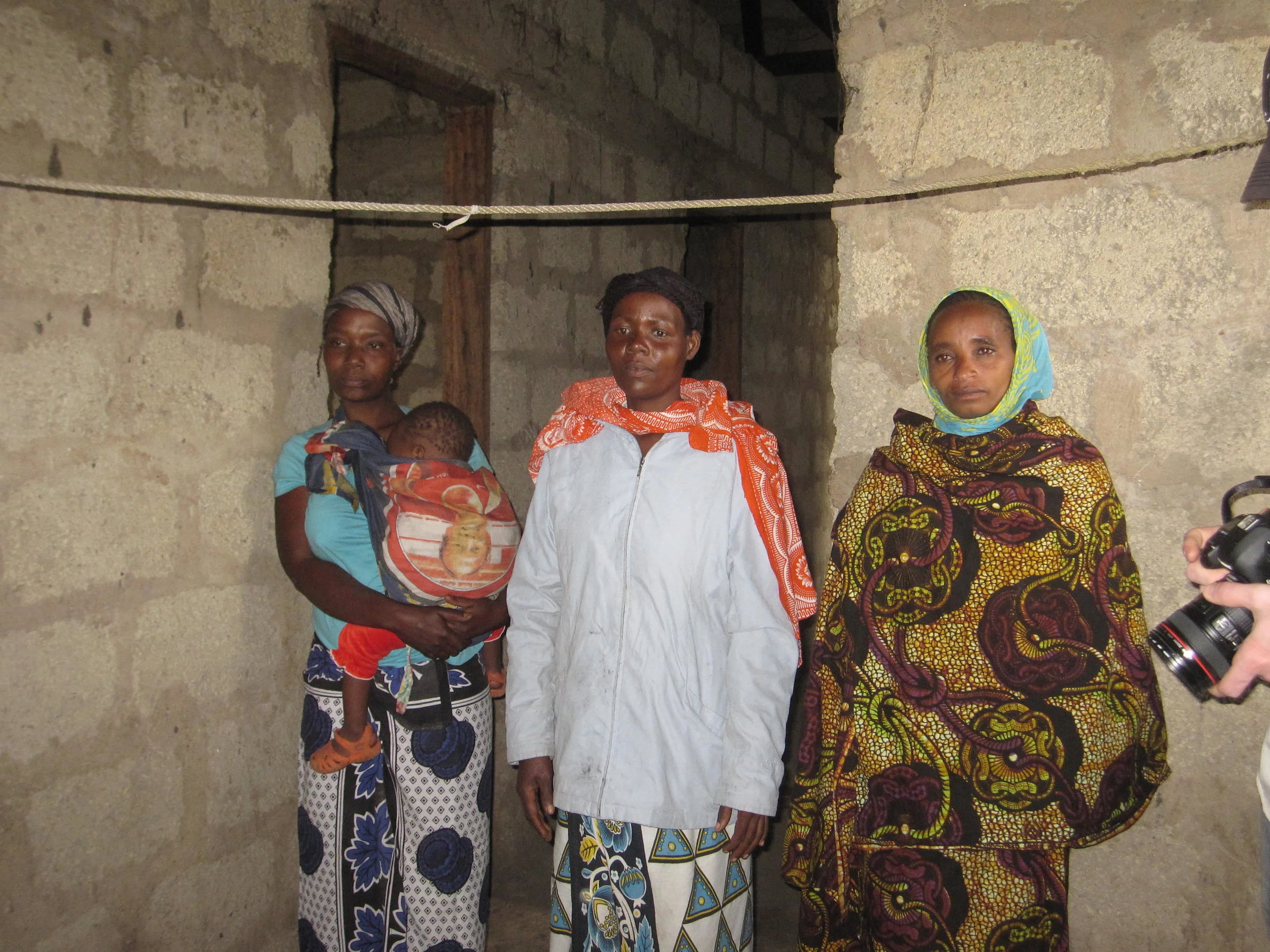 Three women from Tanzania standing inside a concrete room, with one woman holding a child wrapped in a blanket.