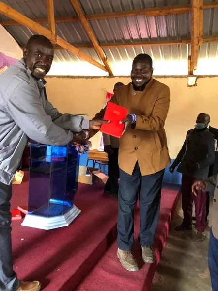 Two pastors are on a stage; one is handing a native translation of a bible to the other while shaking hands. They are smiling and are participating in a ceremony.