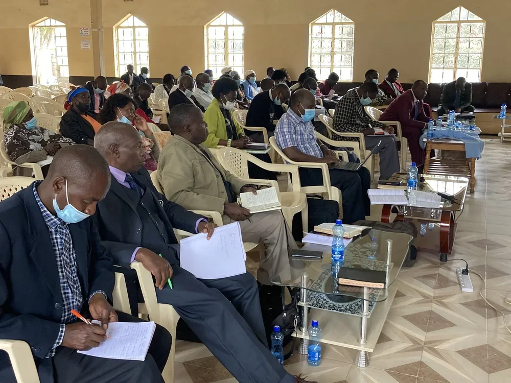 Group of pastors attending a training in a large room, sitting on beige plastic chairs, wearing face masks, with notebooks.