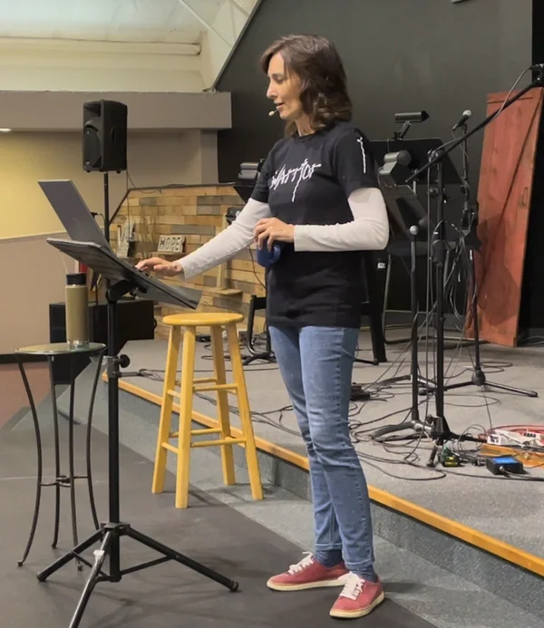Tamara Rowe speaking into a microphone on a stage, with a music stand, a drum set, and microphone stands behind her, in a church or auditorium setting.