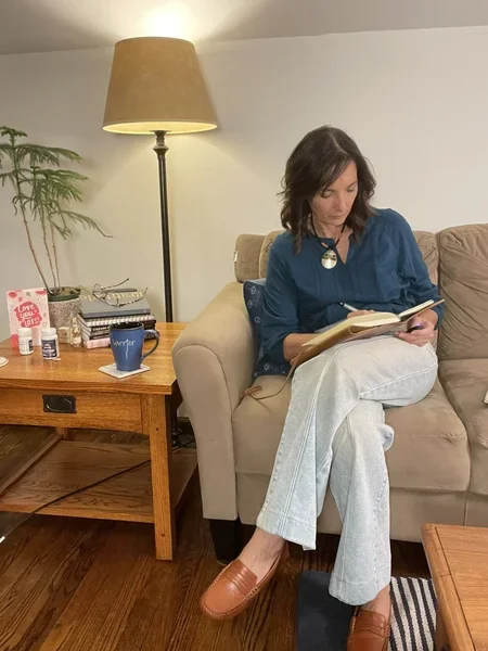 Tamara sitting on a beige sofa reading a book with a black coffee mug labeled 'Warrior' on the table beside her. She is wearing a blue shirt, white pants, and brown shoes. There is a tall floor lamp and various items on the coffee table.