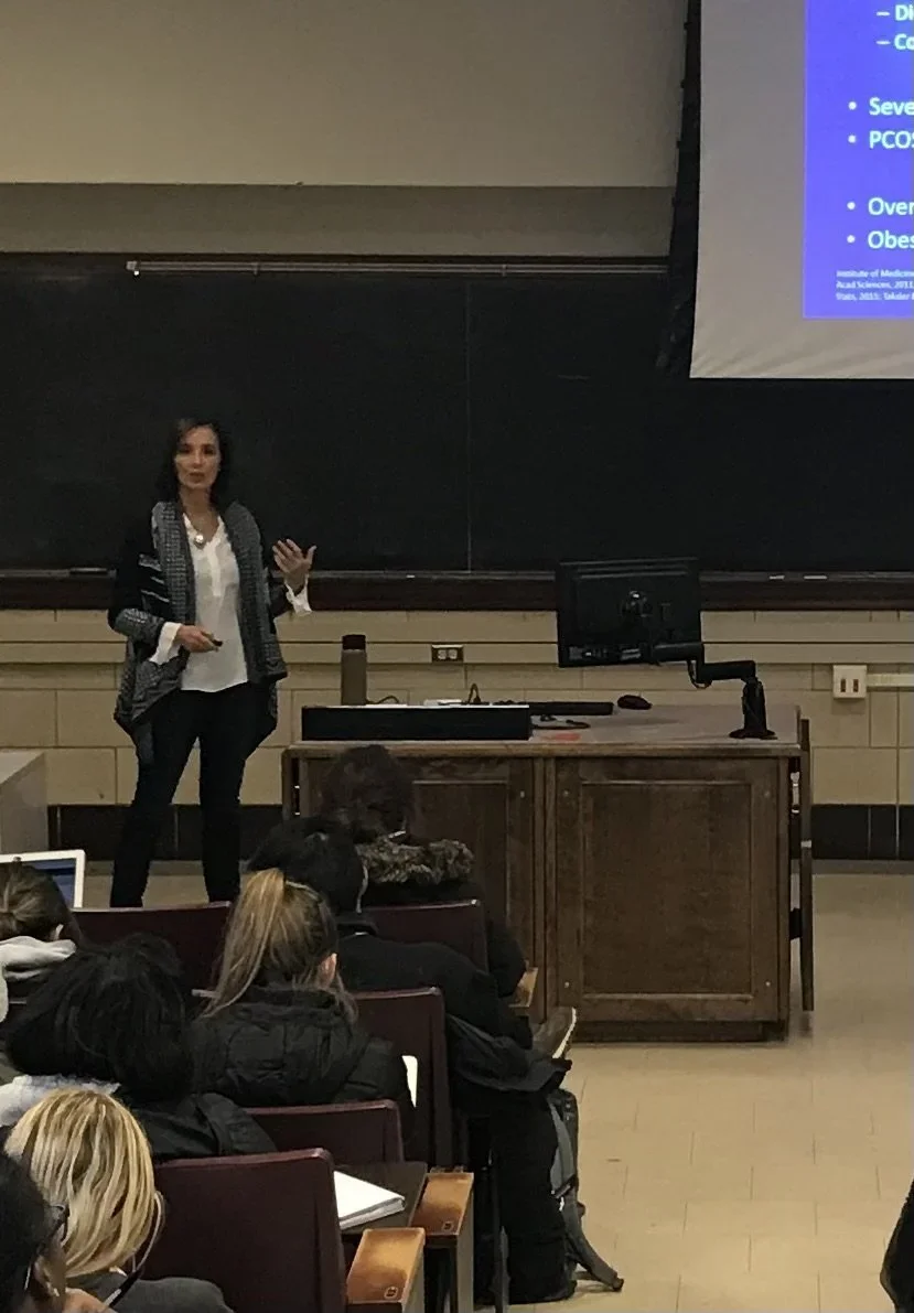Tamara giving a lecture in a classroom with students listening, a blackboard, a projector screen displaying a slide, and a computer on a desk at the University of Illinois on health and wellness.