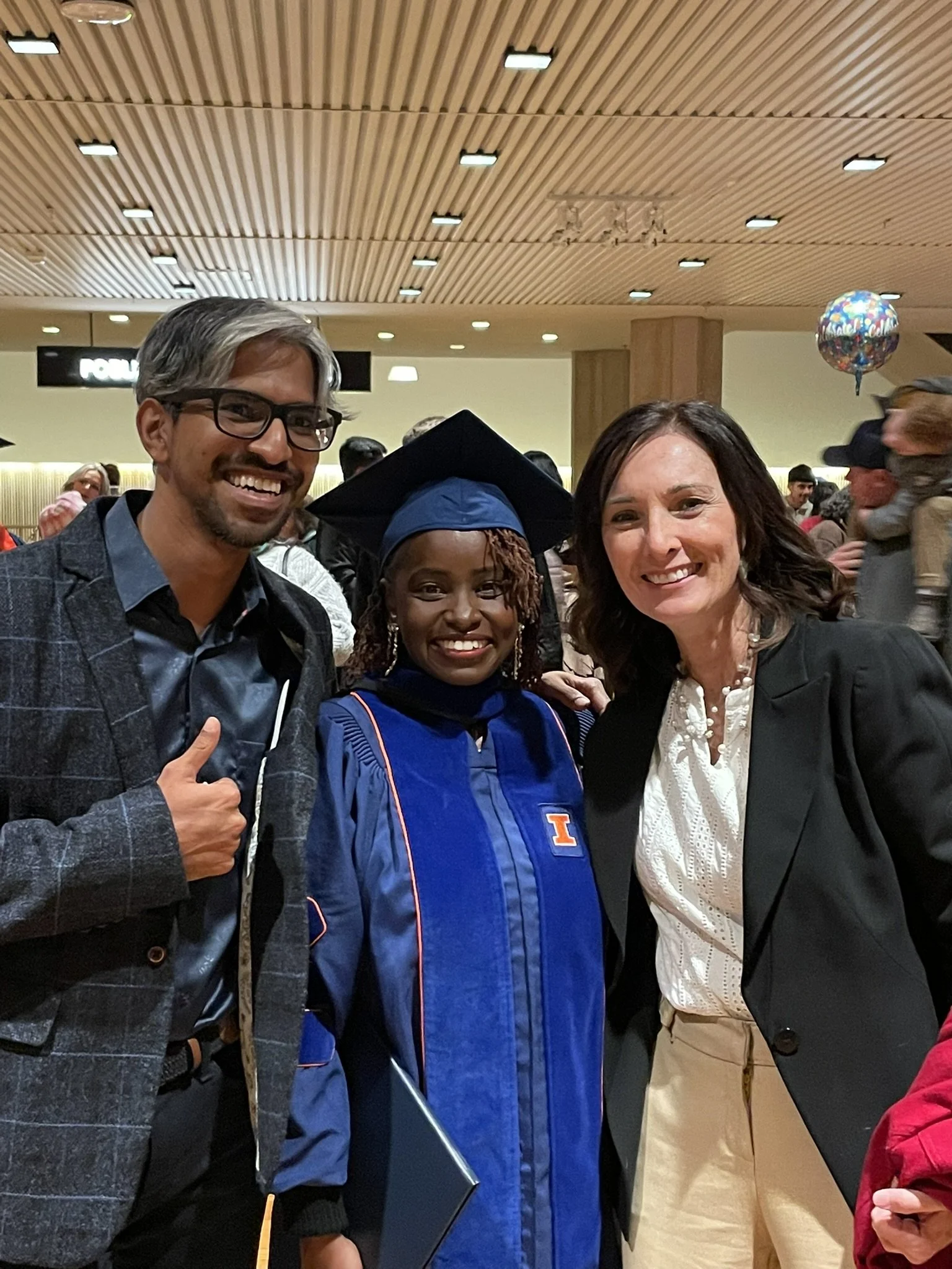 Tamara and two international graduate students smiling, one wearing graduation cap and gown, in a crowded indoor celebration area.