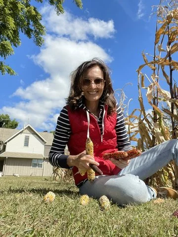 Tamara sitting on the grass in a backyard during fall, smiling and holding two ears of fresh corn.