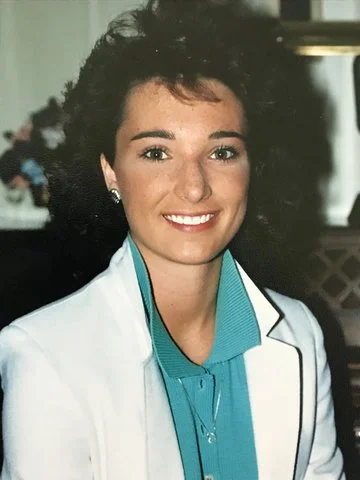 Tamara with short curly dark hair wearing a white coat and blue blouse, indoors in her highschool days.