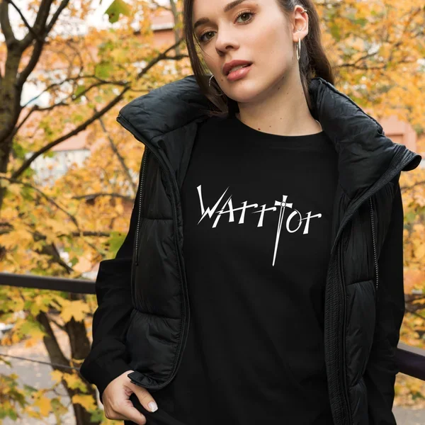 Close-up of a young woman with brown hair,earing a black jacket and the christian warrior graphic T-shirt, standing  woutdoors during autumn with yellow and orange leaves in the background.