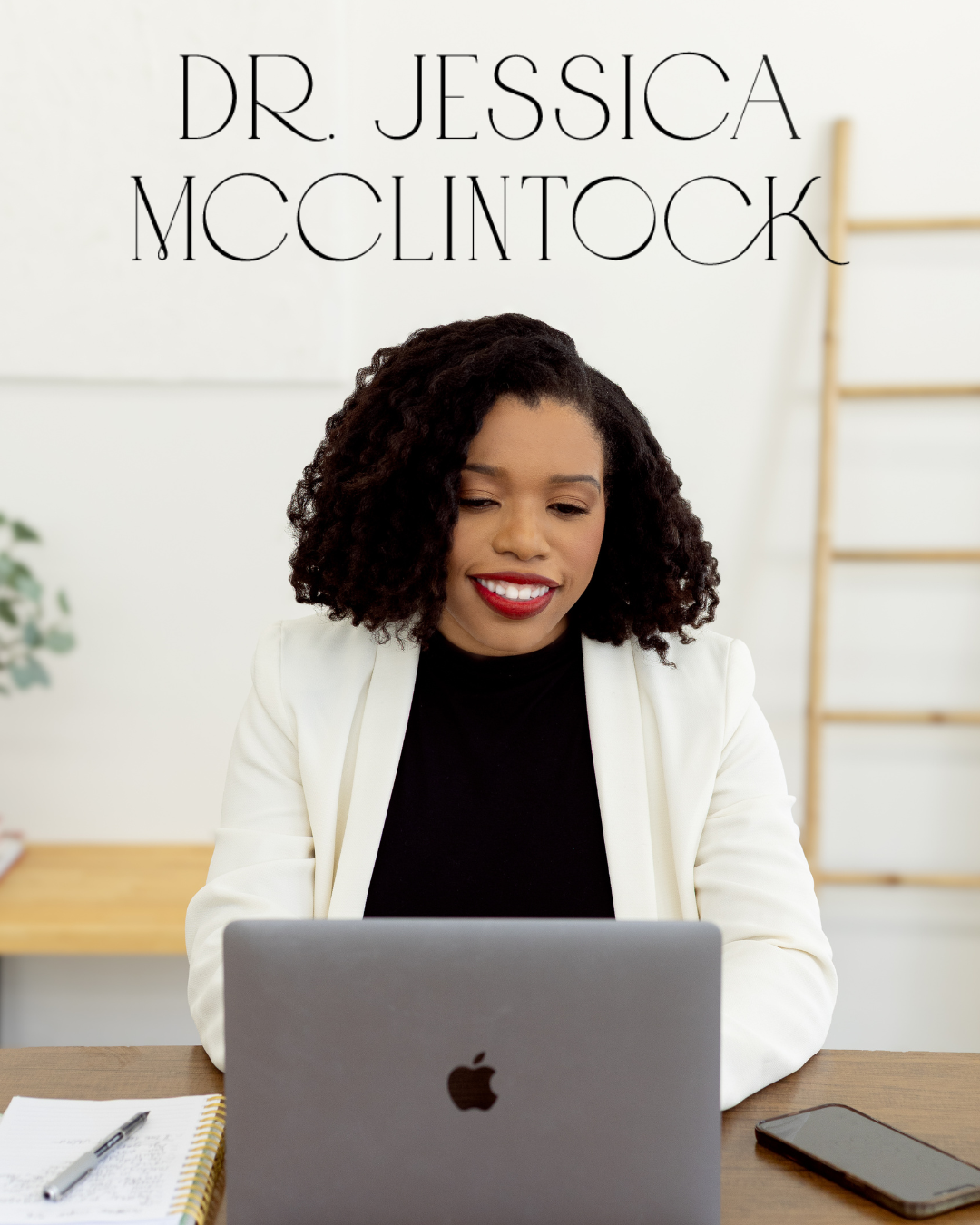 A woman with curly black hair smiling while working on a silver MacBook at a desk.