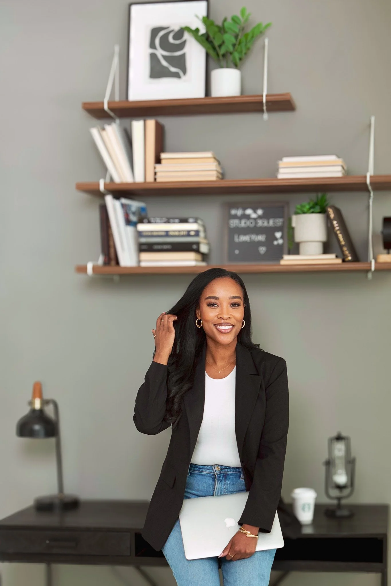 A woman with long dark hair, wearing a black blazer and blue jeans, smiling and holding a silver laptop, standing in a modern office space with a green accent wall, bookshelves, and decorative items in the background.