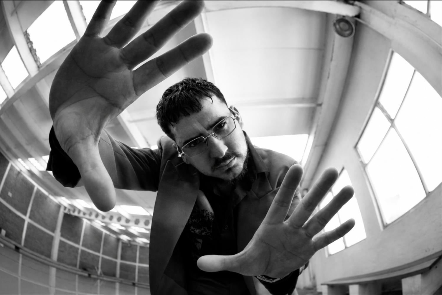 A man with glasses reaching out towards the camera with both hands, looking directly at it, in an industrial-style building with large windows.