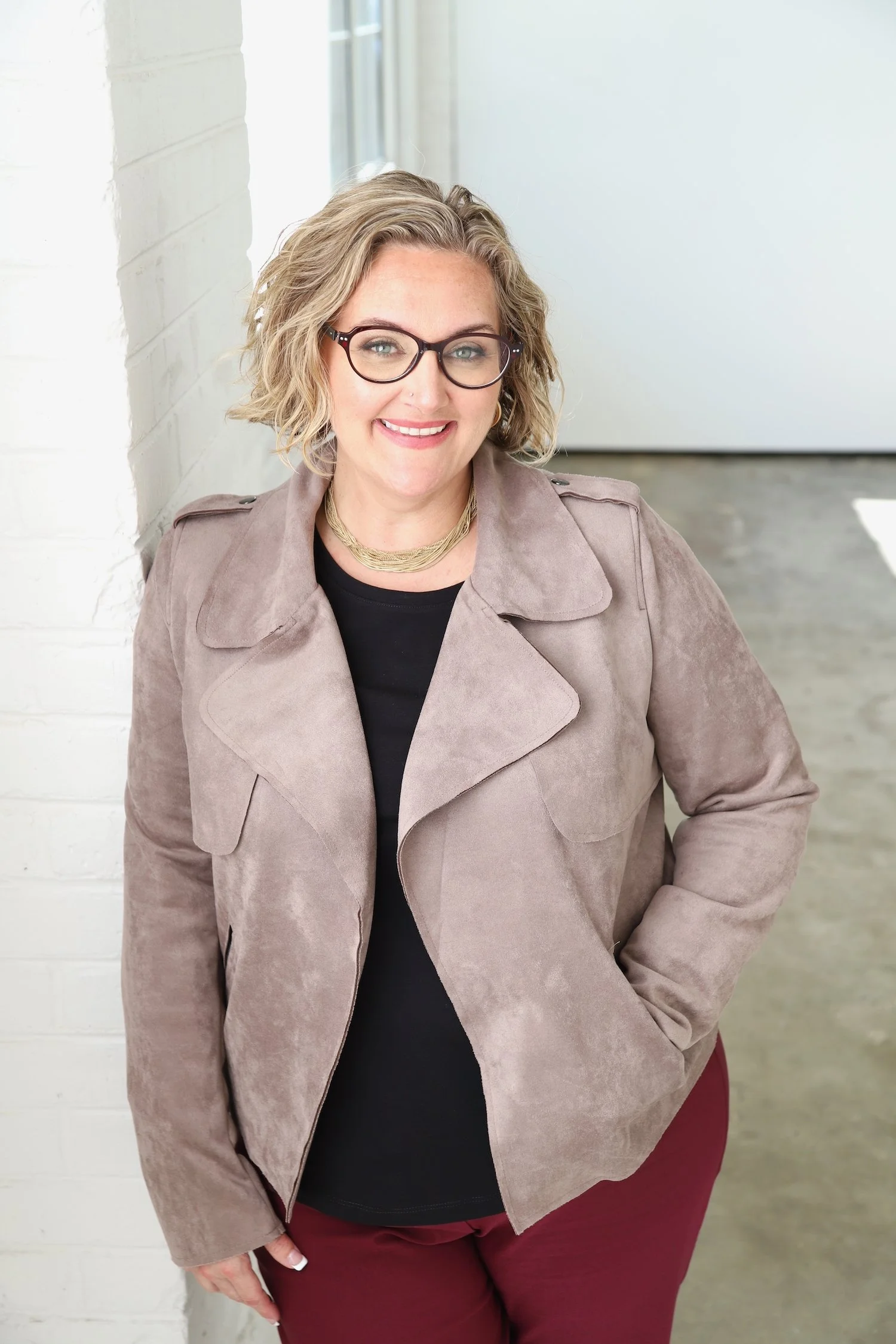 Jess Sato in a black top, tan jacket, and maroon pants leaning against a wall