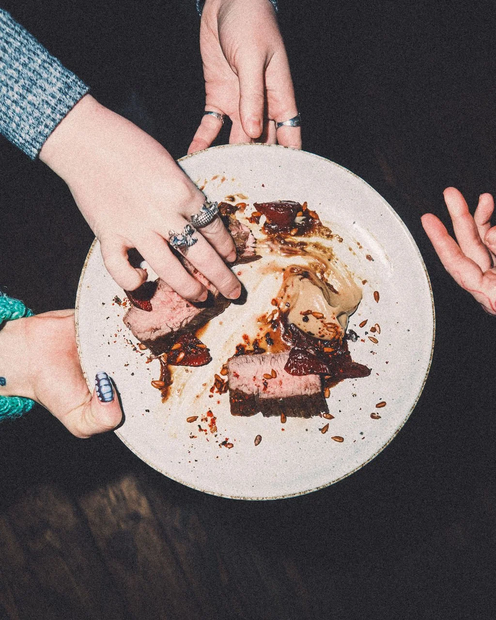 Hands reaching for a plate of sliced beef, vegetables, and sauce on a white plate.