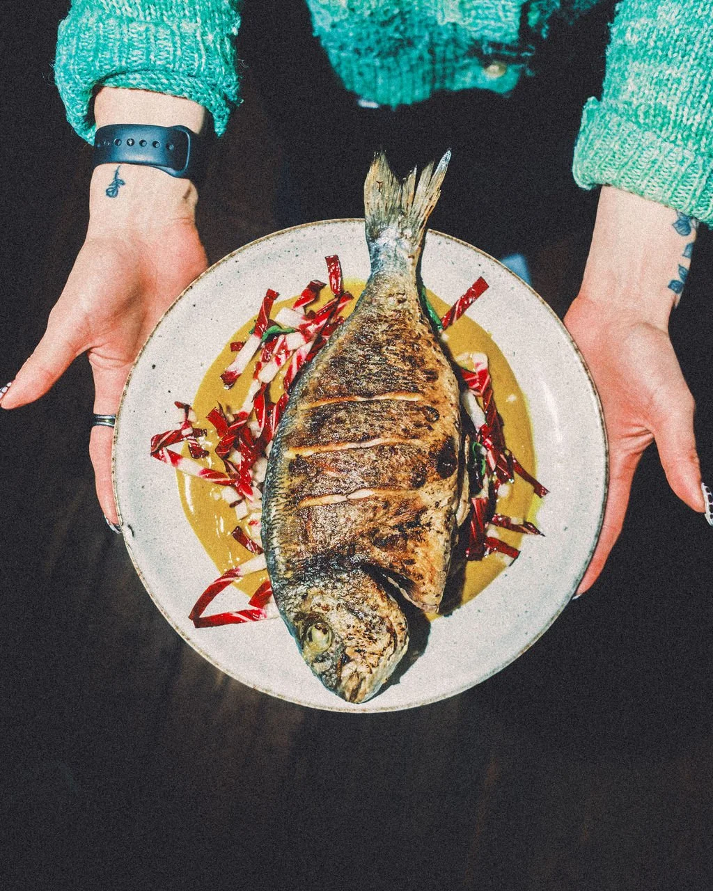 A person holding a plate with a whole grilled fish garnished with red and white vegetable strips.
