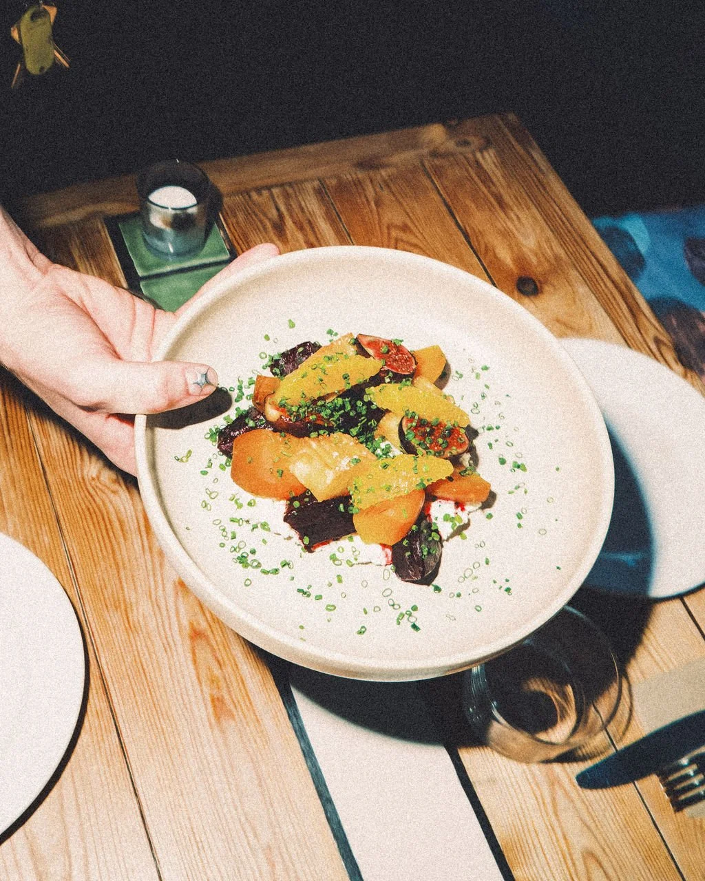 Person holding a plate of colorful sliced vegetables, garnished with chopped green herbs, on a wooden table with a small candle in the background.