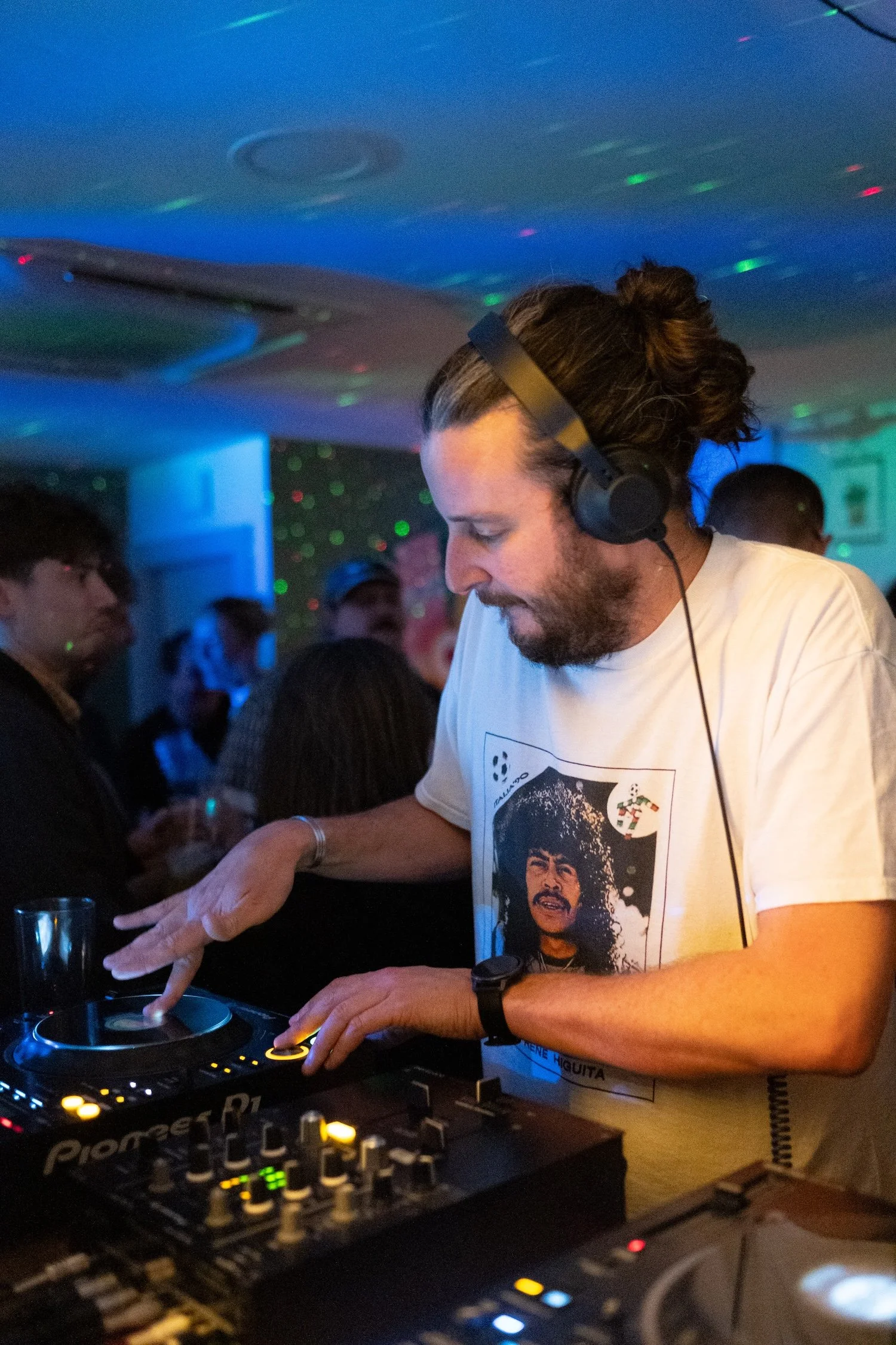 A man with long hair tied in a bun, wearing a white T-shirt with a graphic print, DJing at a party with colorful lights and other attendees in the background.