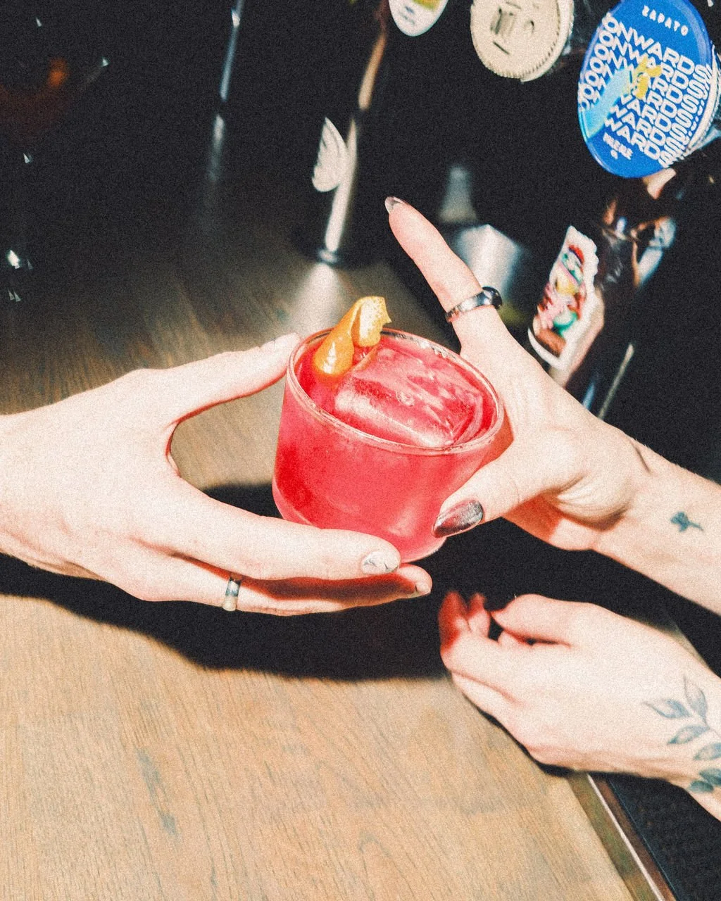 Two hands holding a red cocktail glass with an orange wedge garnish, set on a dark wooden table.