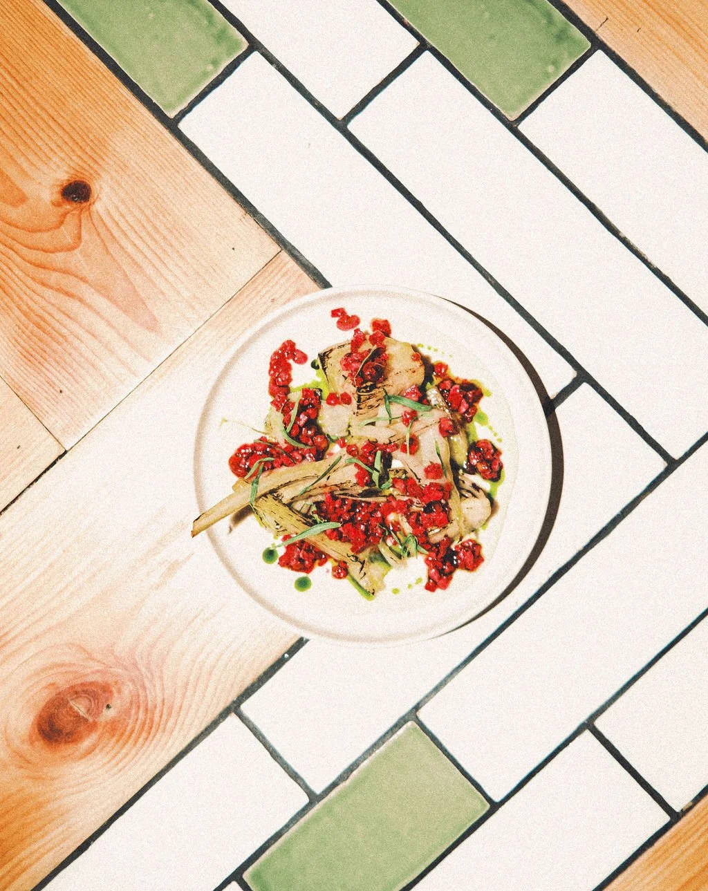 A plated dish with grilled vegetables topped with chopped red peppers and herbs, garnished with green oil drizzle, on a white plate on a tiled table.