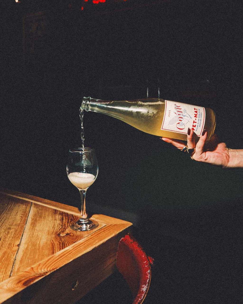 A person pouring sparkling wine or champagne from a bottle labeled Cava Pet-Nat into a wine glass on a wooden table with a dark background.