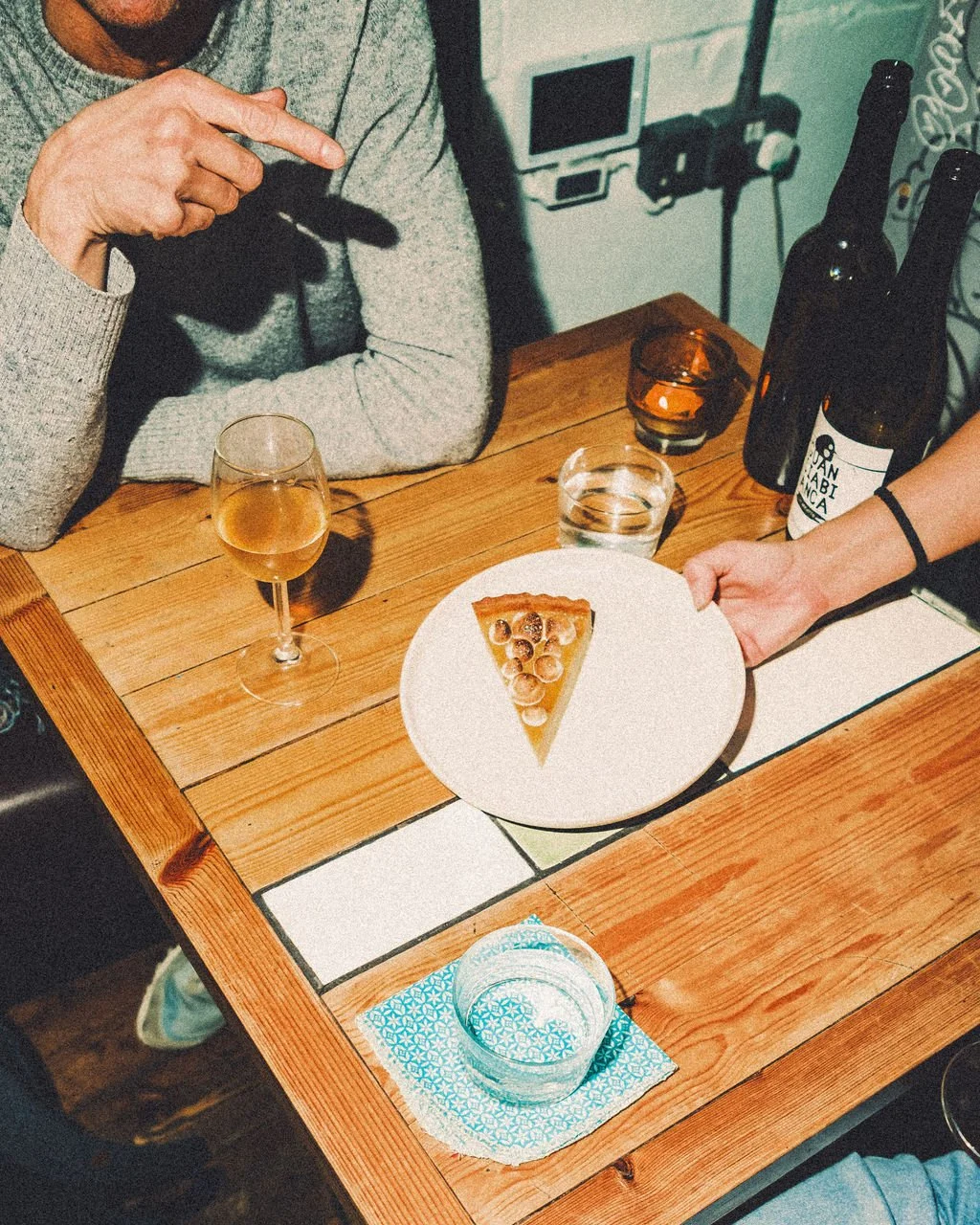 A restaurant setting with a person sitting at a wooden table, receiving a slice of pizza on a white plate from a server. The table has a glass of white wine, a glass of water, a candle, and bottles of wine.